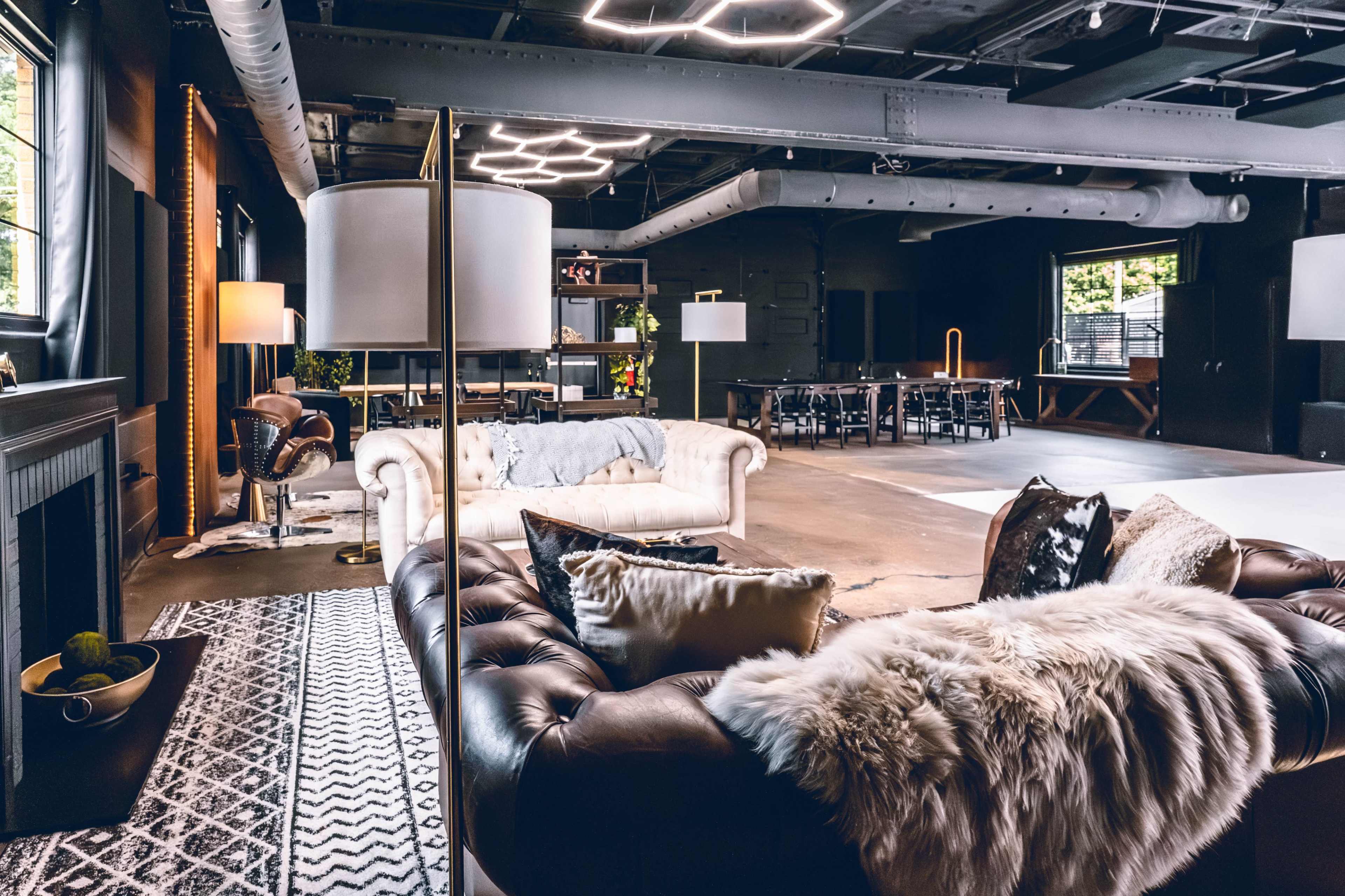 The image shows a modern, spacious lounge area featuring a leather couch, a patterned rug, and various seating arrangements, all within an industrial-style room with exposed ceilings and beams.