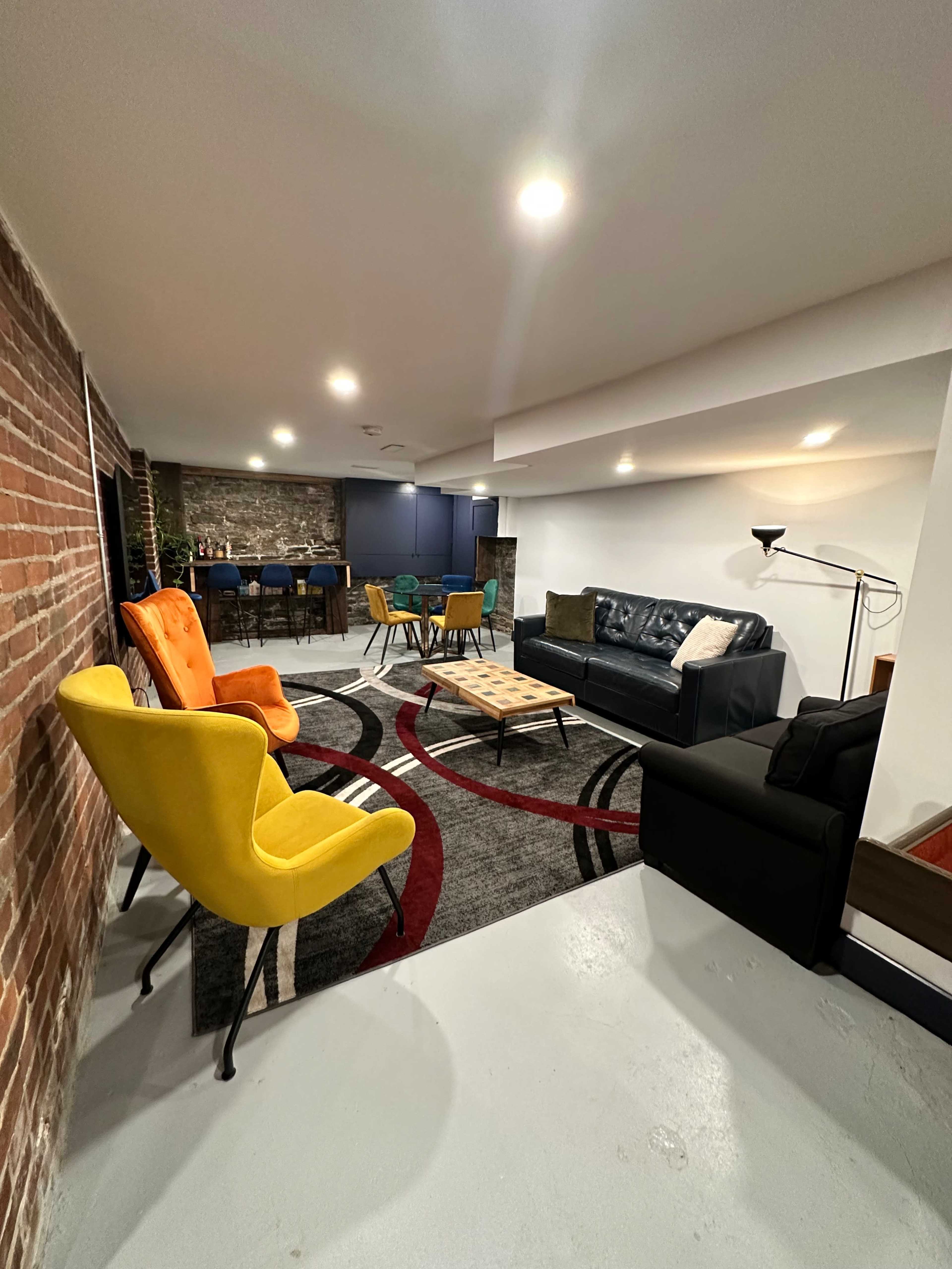 A cozy basement lounge area featuring colorful armchairs, a dark sofa, a coffee table, and exposed brick and stone walls.