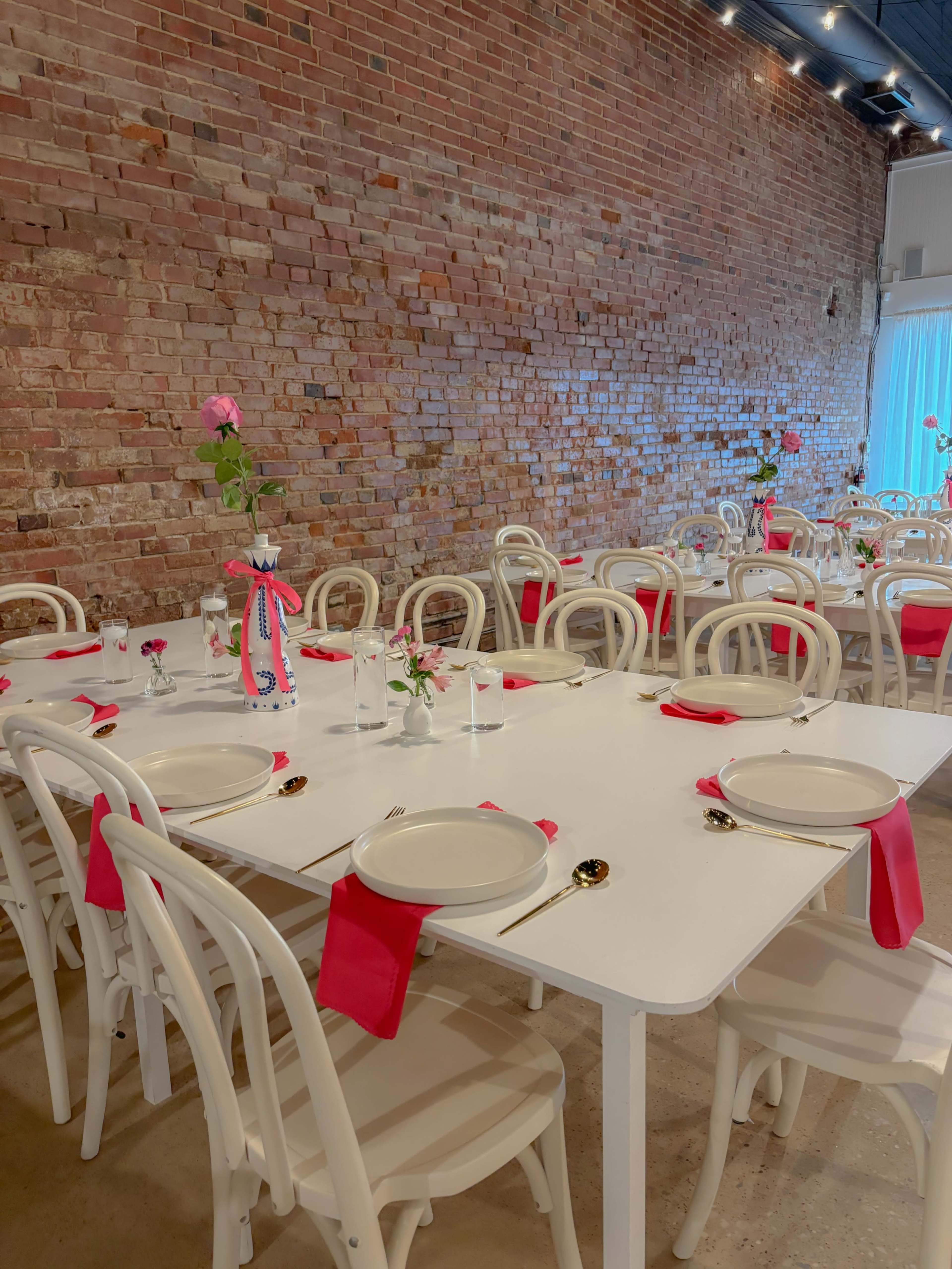 A dining area featuring white tables and chairs, adorned with red napkins and small flower vases, set against a brick wall.