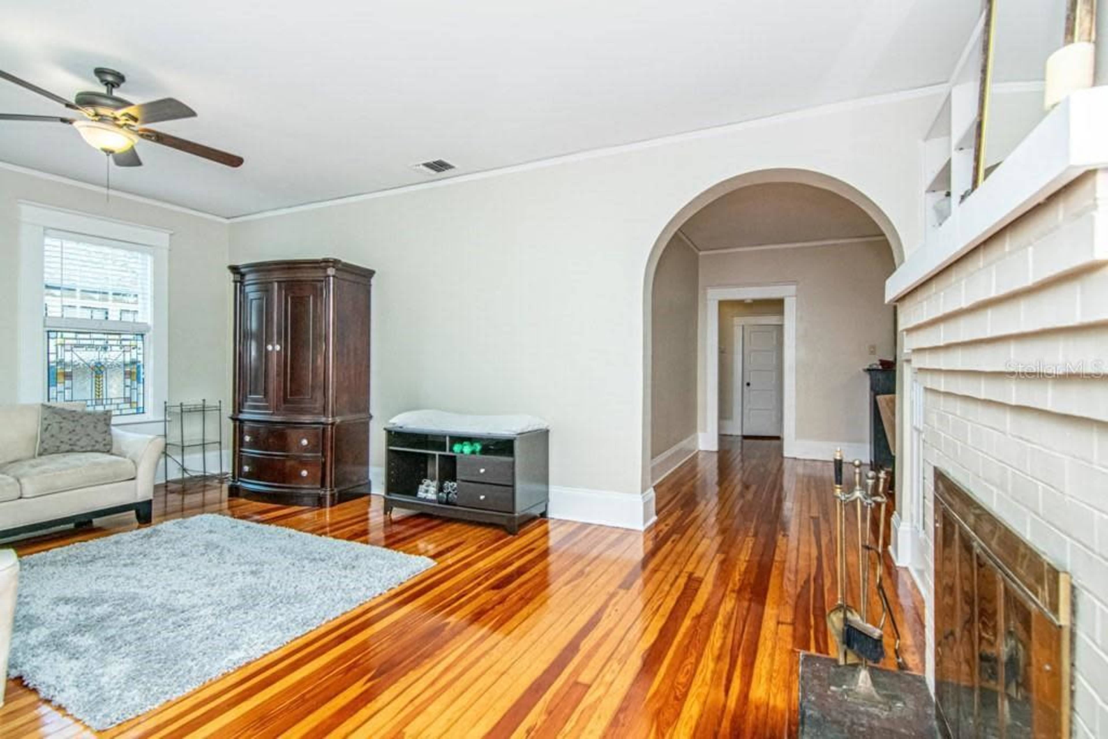 The image shows a living room with polished wooden floors, a cream-colored couch, a dark wooden cabinet, and an arched entryway leading to another room.