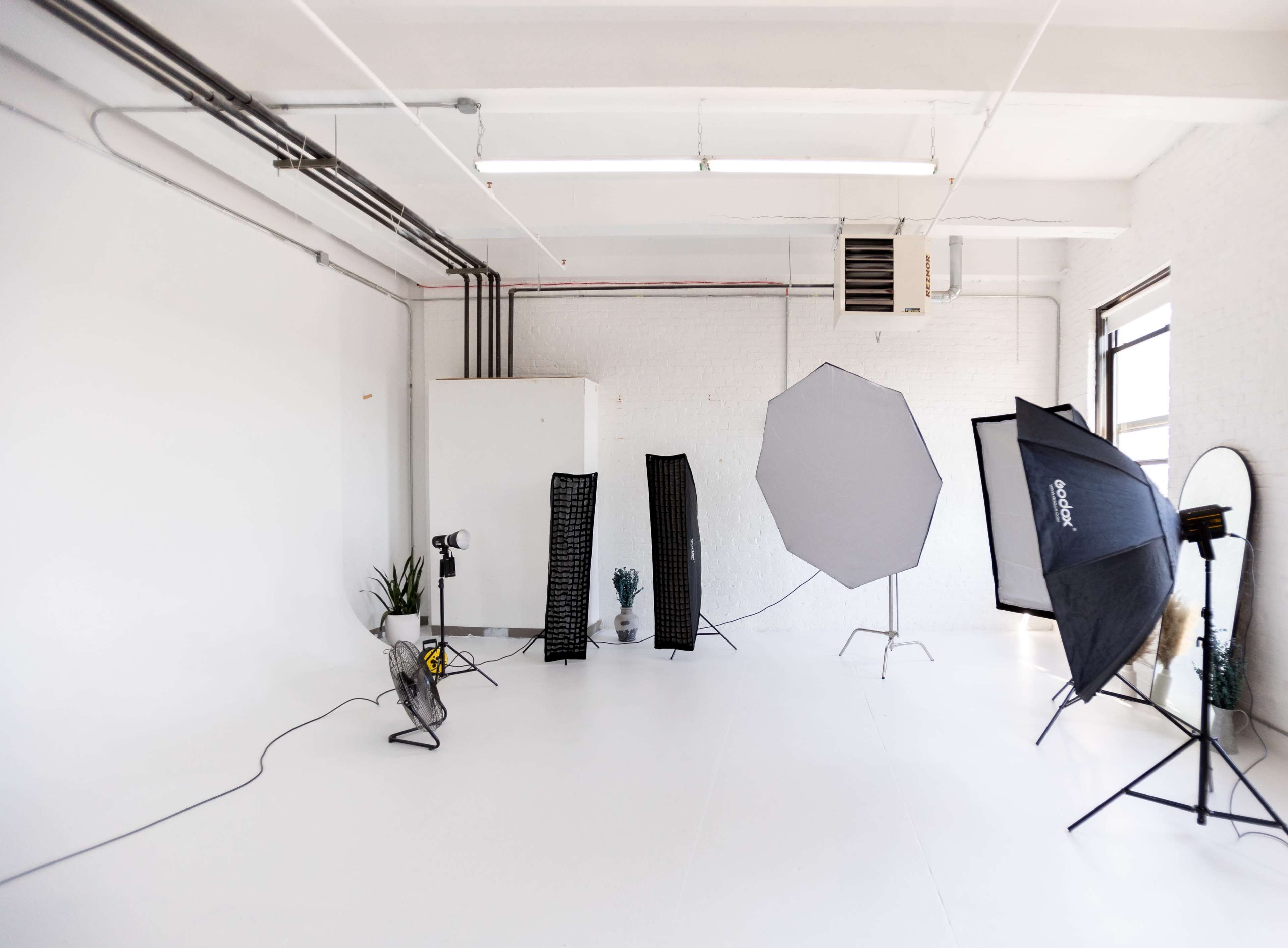 A photography studio with a clean, white backdrop, featuring various lighting equipment and props arranged around the space.
