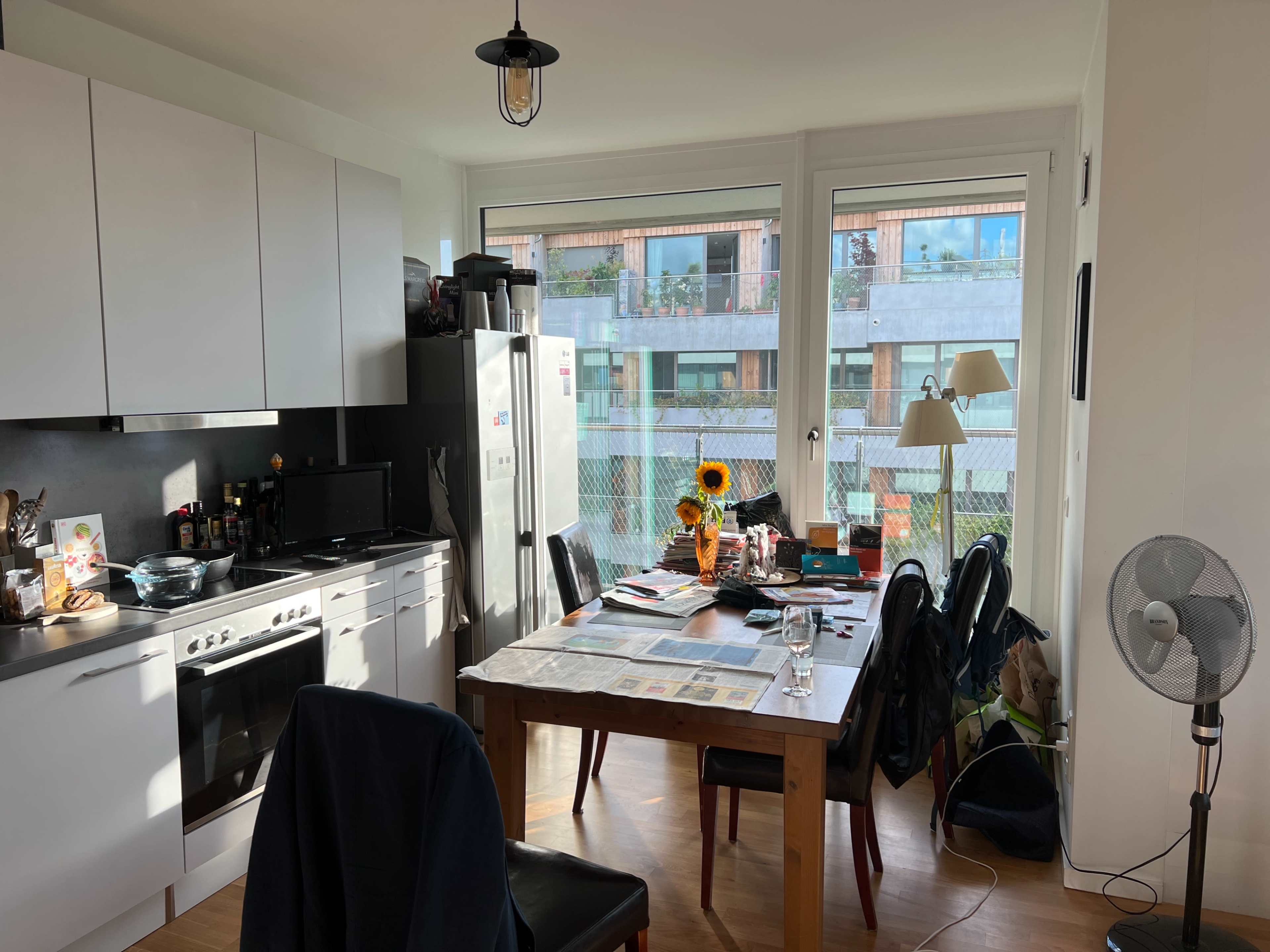 A kitchen-dining area features a table with a sunflower arrangement, chairs, various kitchen appliances, and large windows overlooking an outdoor space.