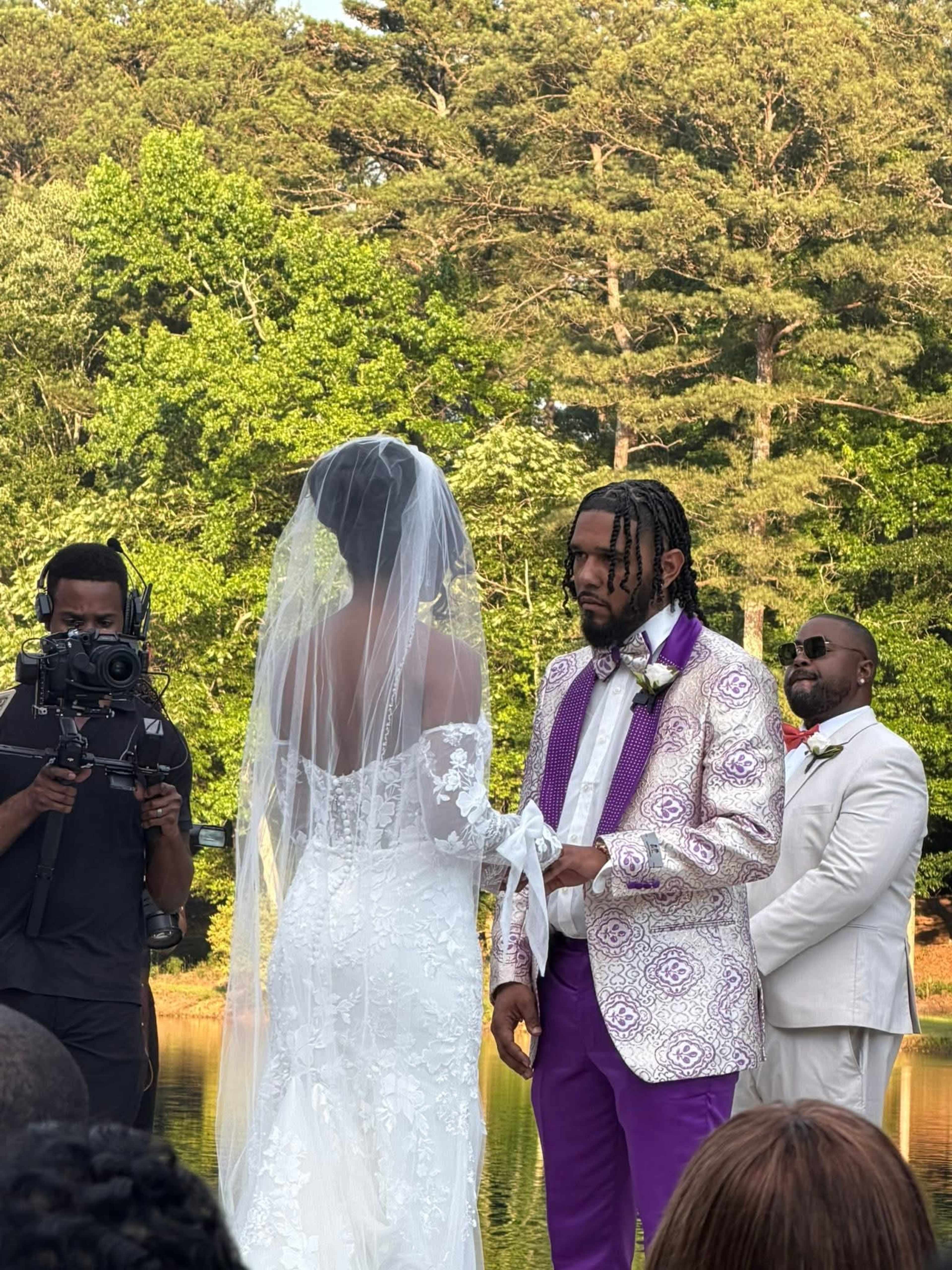 A couple exchanges vows by a body of water while a videographer captures the ceremony and a man in a suit observes.