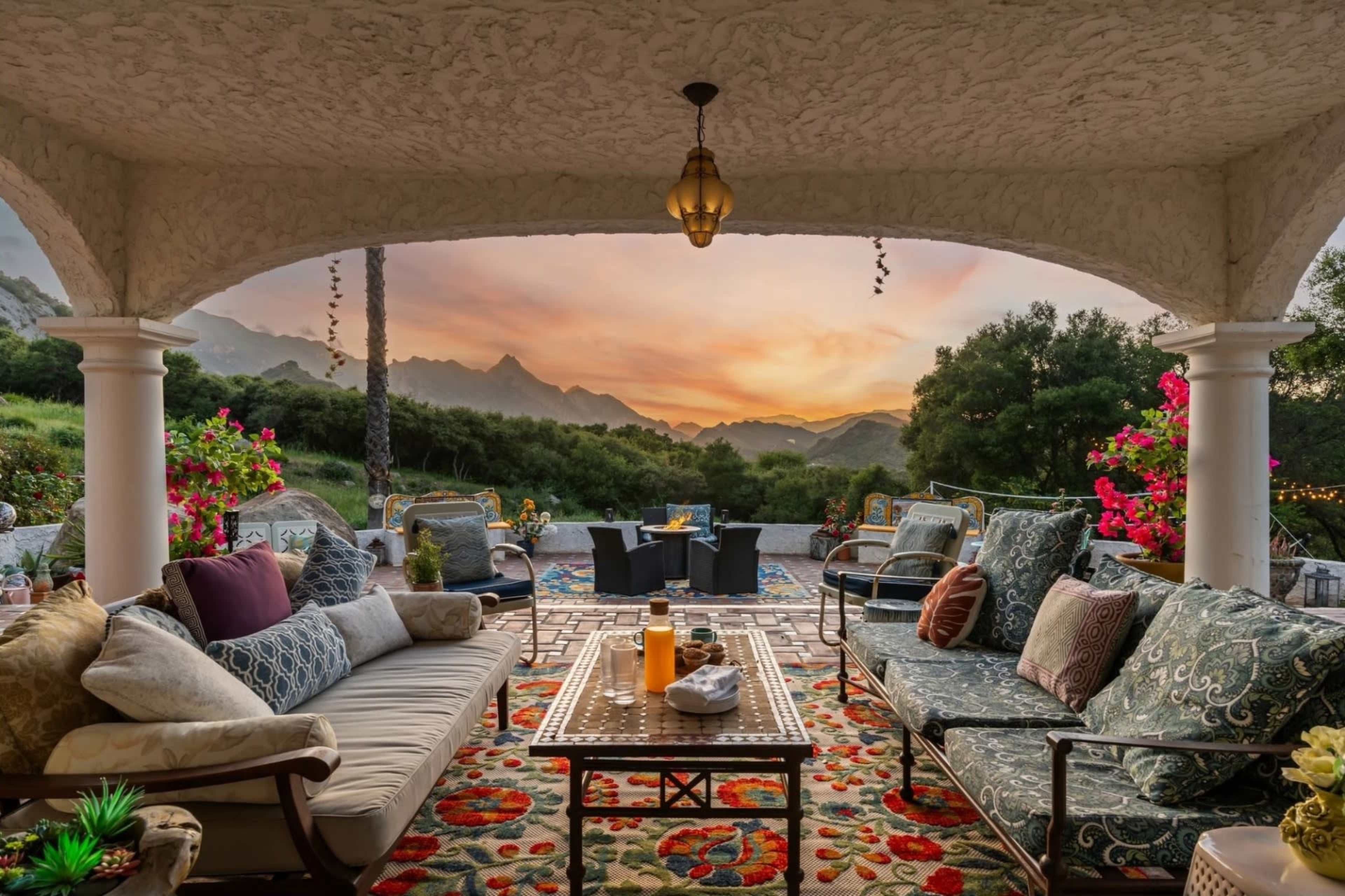 A covered patio features ornate furniture with colorful cushions, a decorative rug, and a scenic view of mountains at sunset.