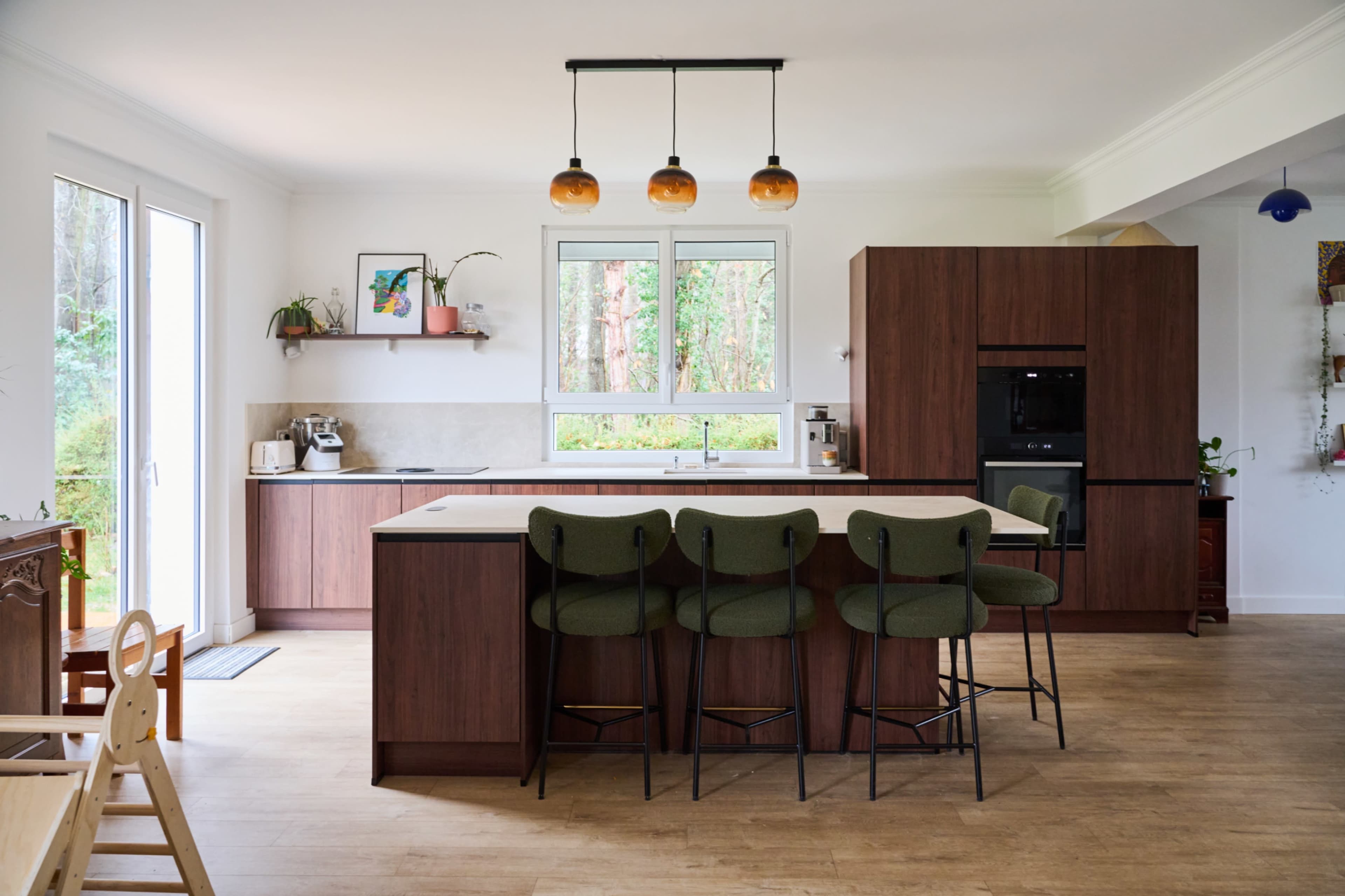 The image shows a modern kitchen featuring a large island with green bar stools, wooden cabinetry, and large windows overlooking a garden.