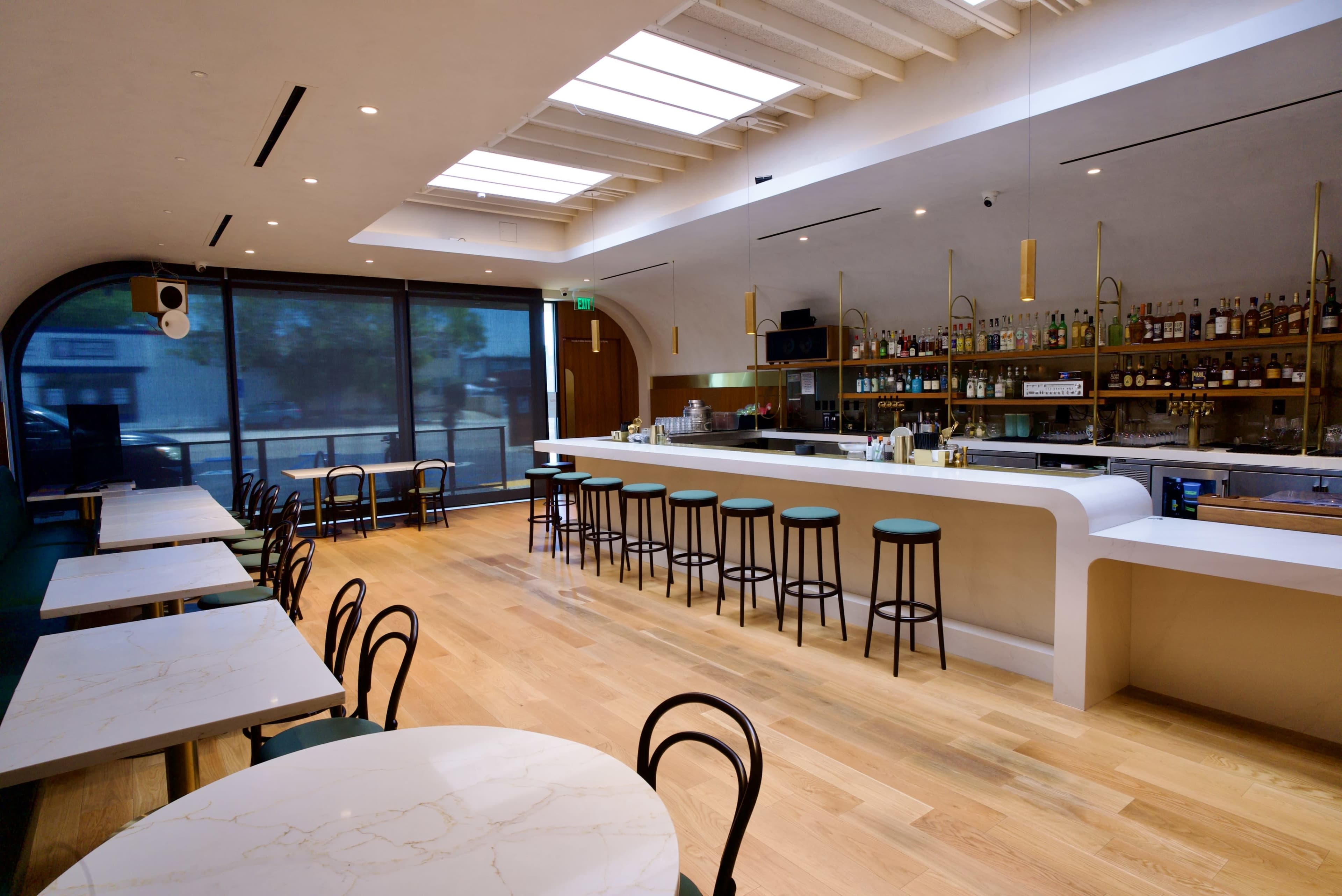 A modern bar area features a white countertop with high stools, surrounded by tables and large windows that let in natural light.