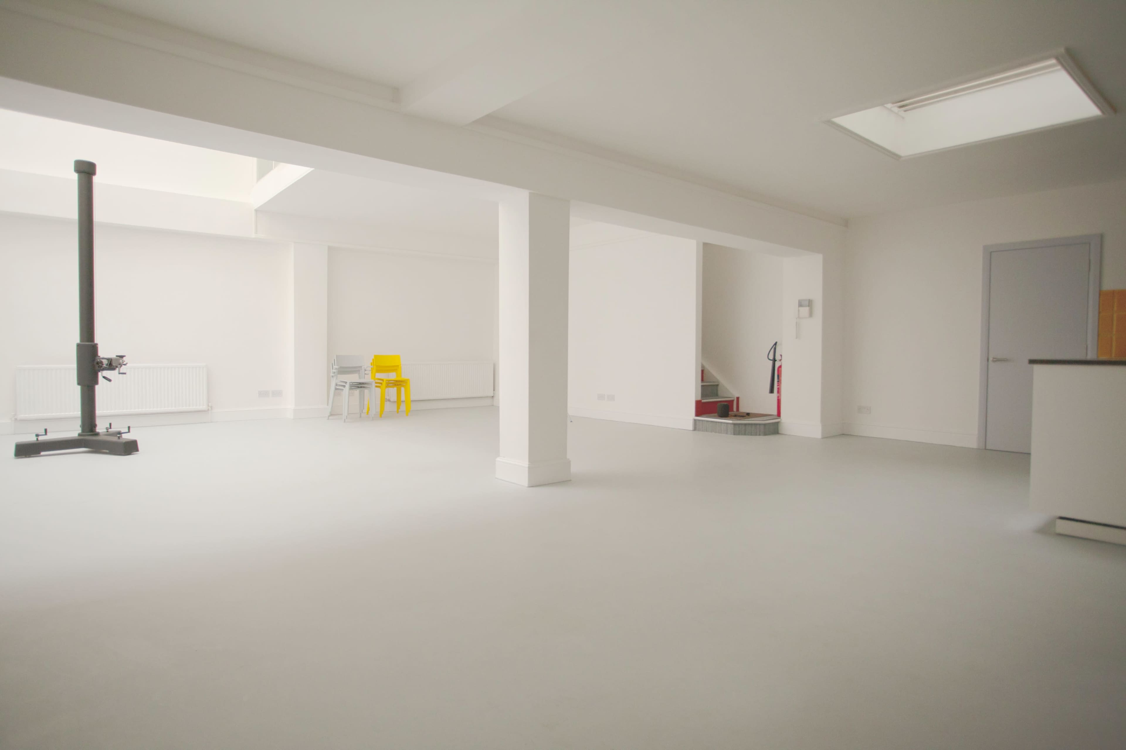 A spacious, minimalistic interior with white walls, a gray floor, and a skylight, featuring a staircase, a fire extinguisher, and a few chairs.
