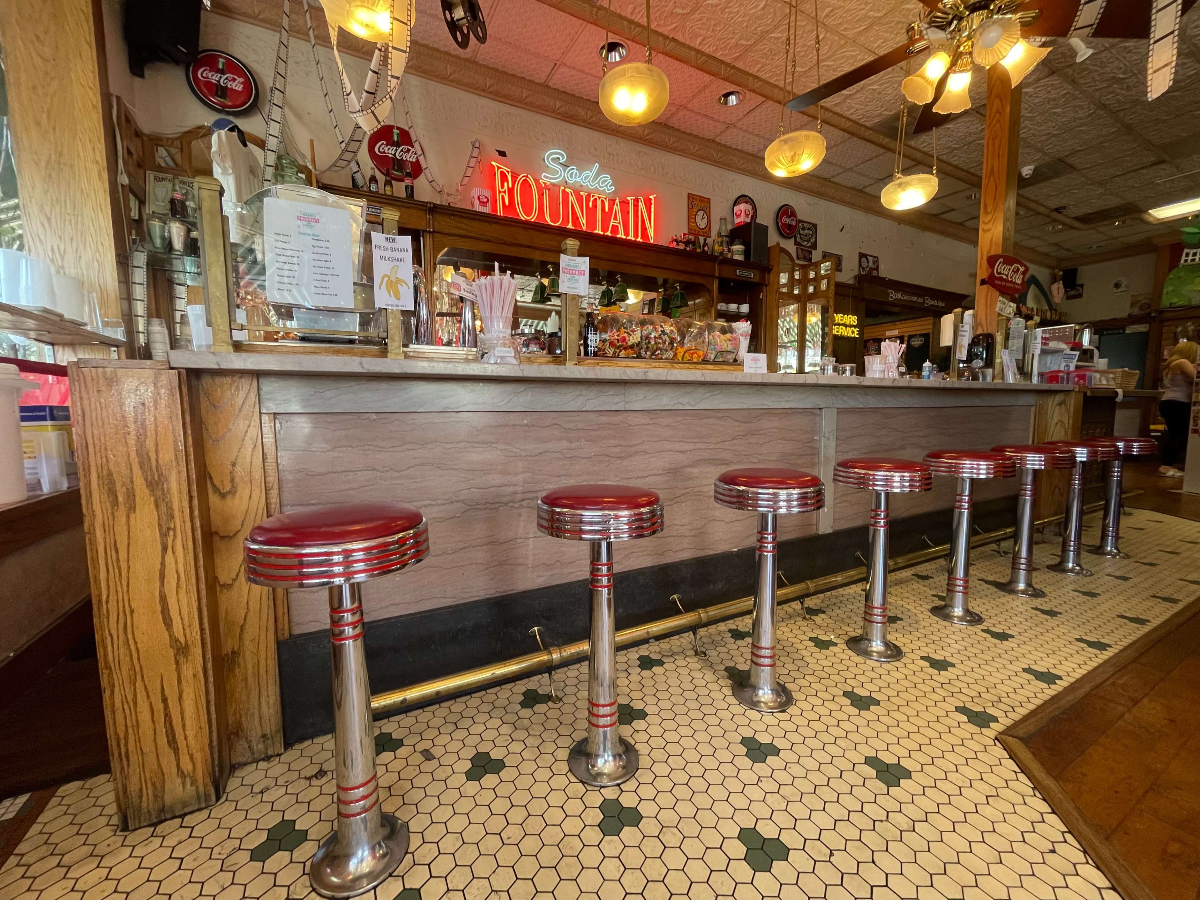 A vintage soda fountain counter with red and chrome stools lined up in front, featuring a menu and display of candy and drinks.