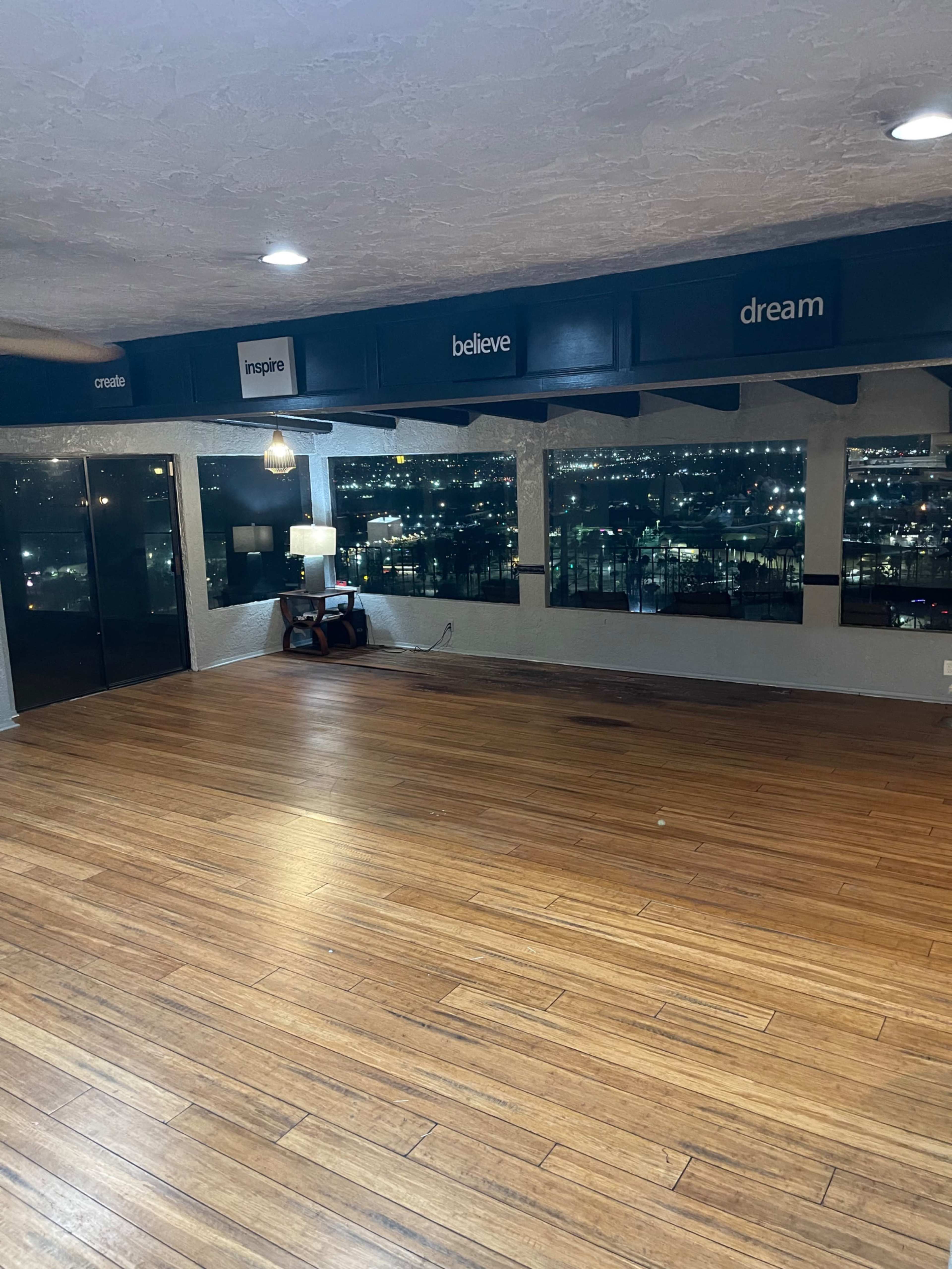 A spacious room with wooden flooring, large windows revealing a city view, and motivational words on a decorative beam.