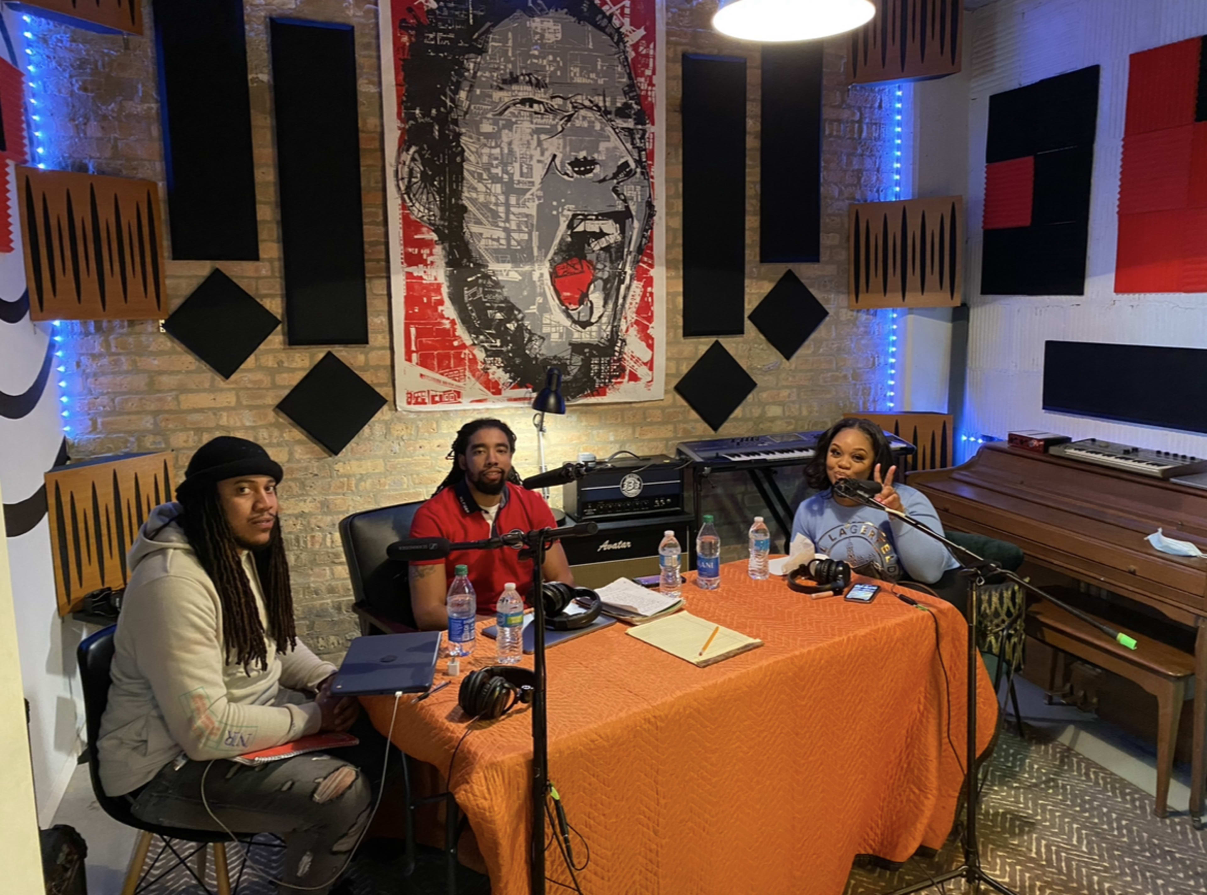 Three individuals are seated around a table with recording equipment in a studio decorated with a large portrait and sound panels.
