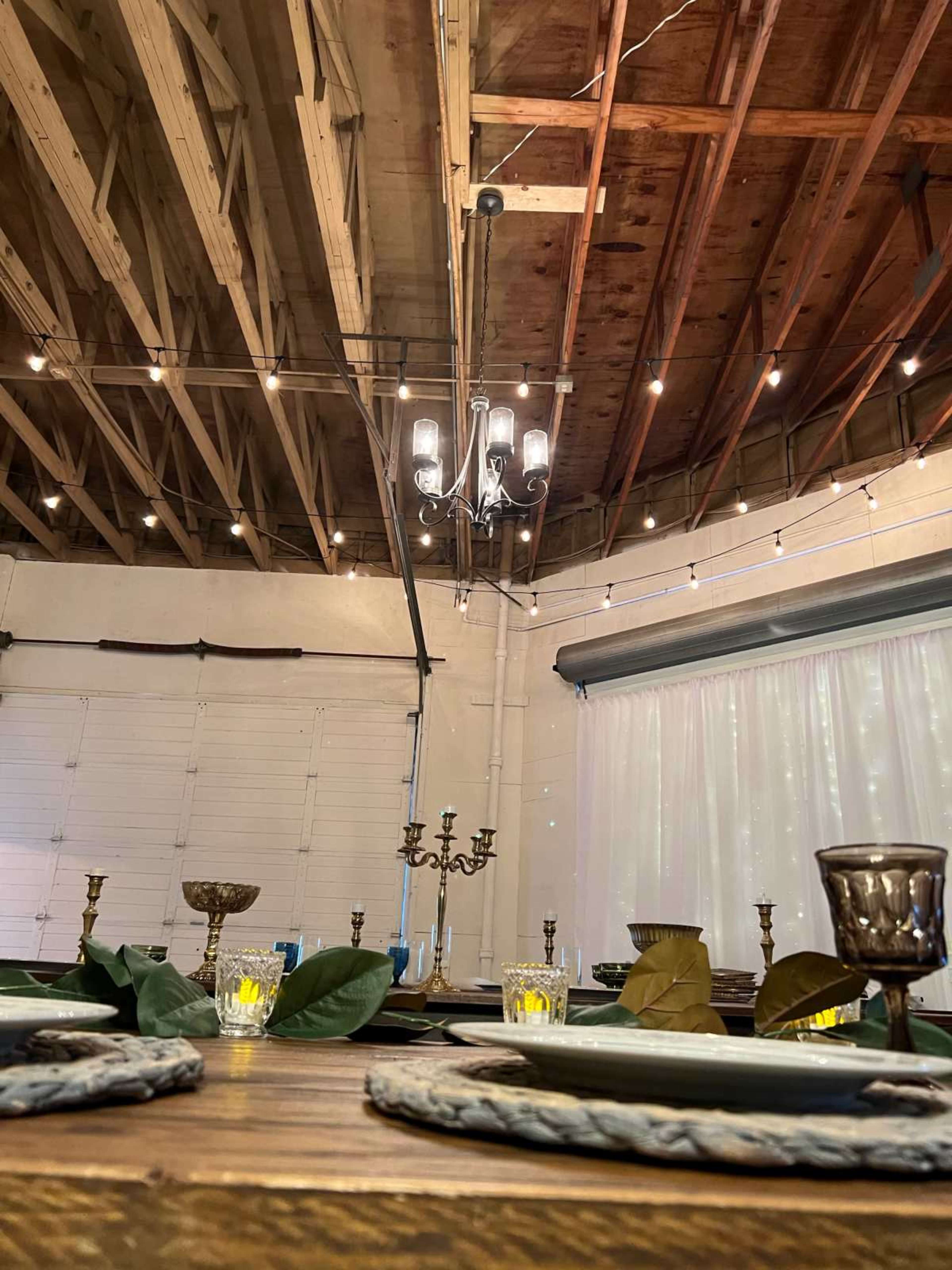 A rustic dining setup with wooden tables, elegant candle holders, and draped white fabric in a warmly lit venue featuring exposed beams and string lights overhead.