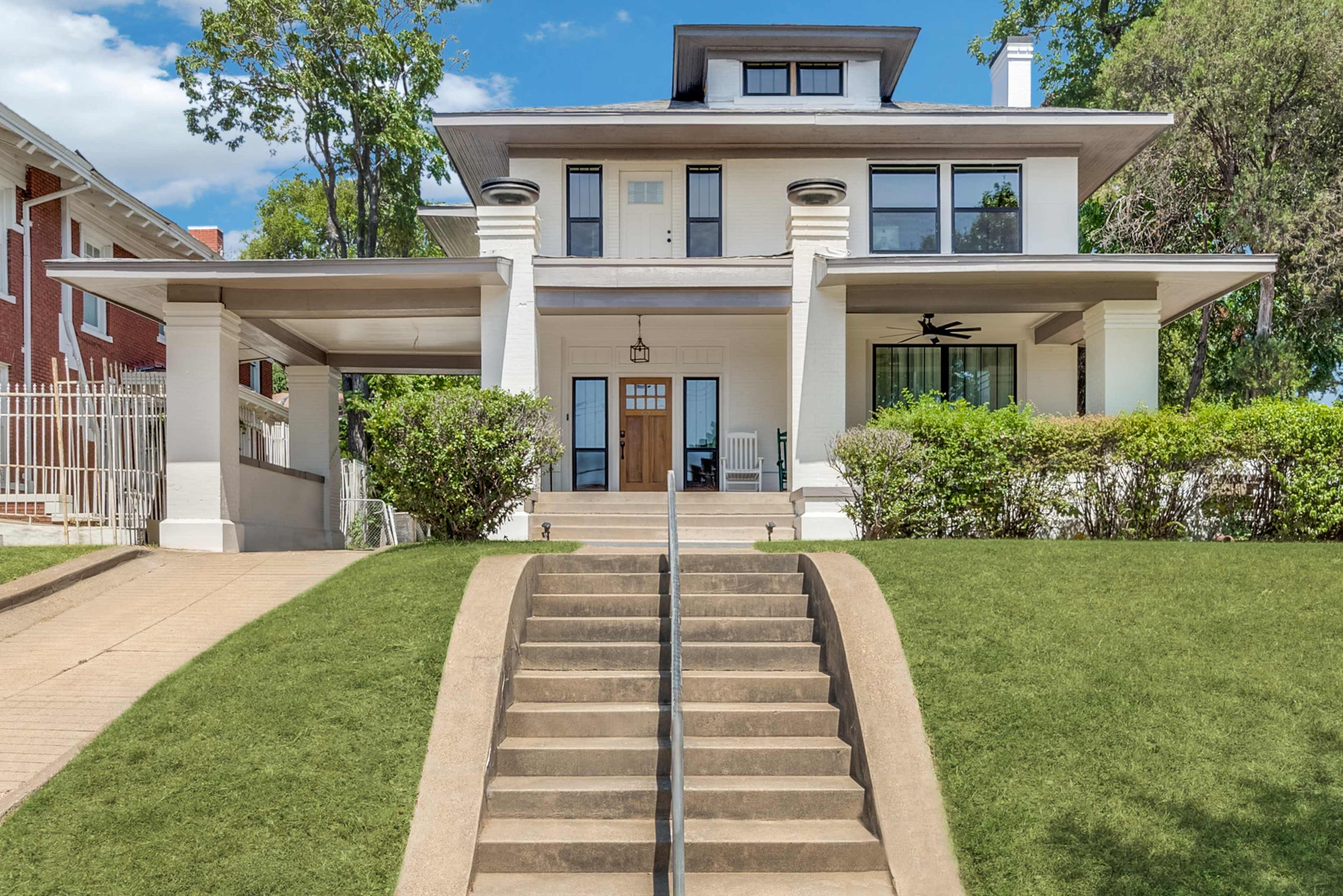 The image shows a three-story house with a large front porch and a set of stairs leading up to the entrance, surrounded by greenery.