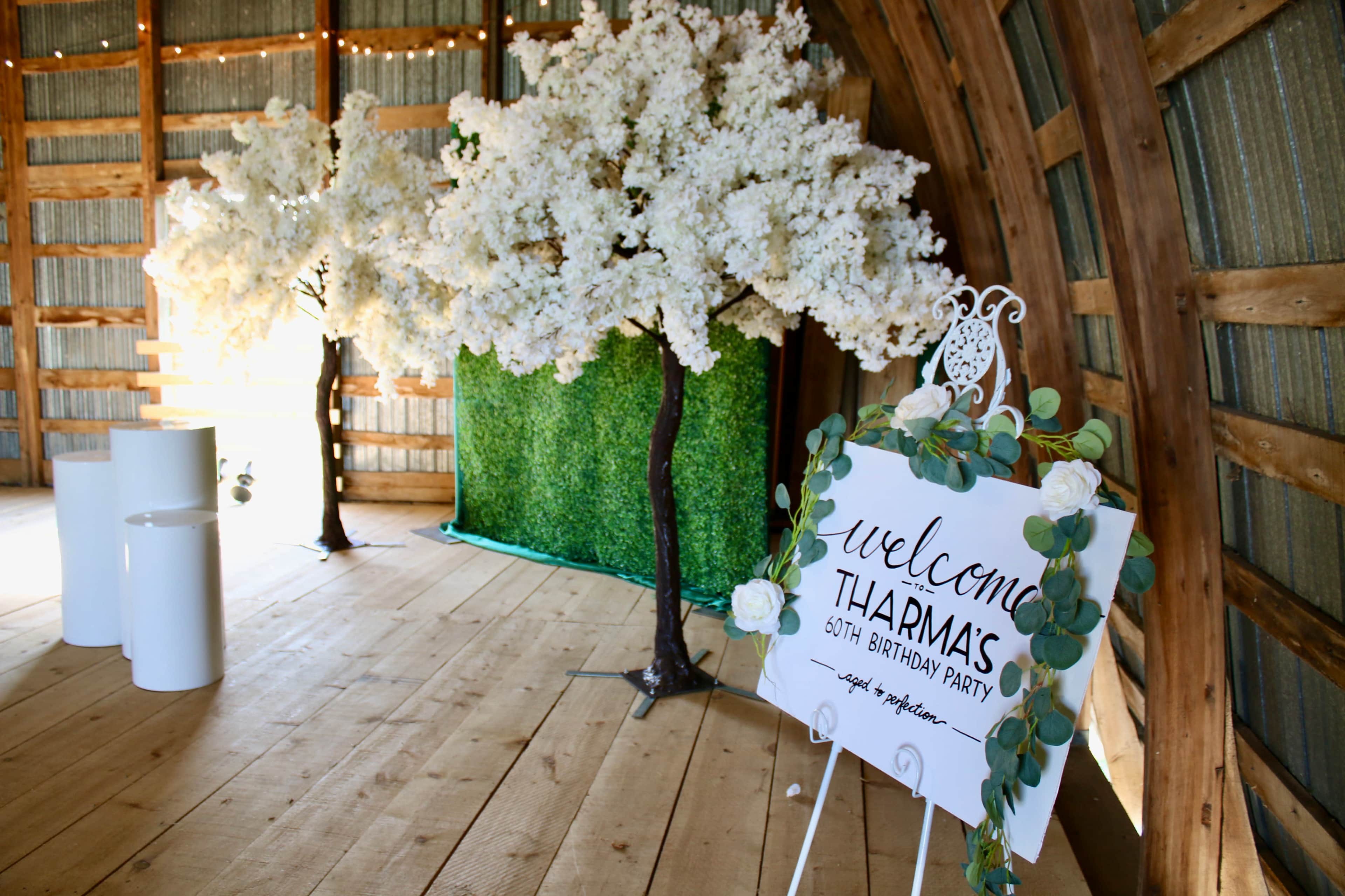 The scene shows a decorated barn interior with white flowering trees, a green backdrop, and a welcome sign for a 60th birthday party.