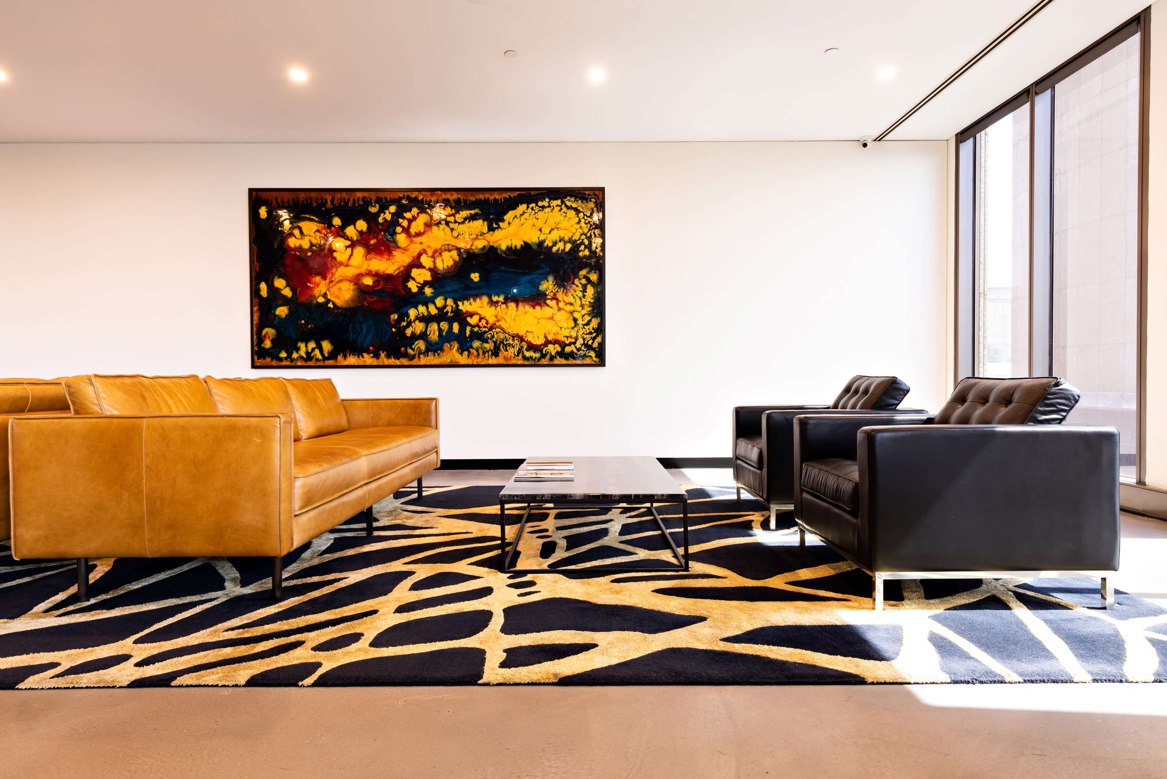 The image shows a modern lounge area with a brown leather sofa, black armchairs, a coffee table, and a vibrant abstract painting on the wall.