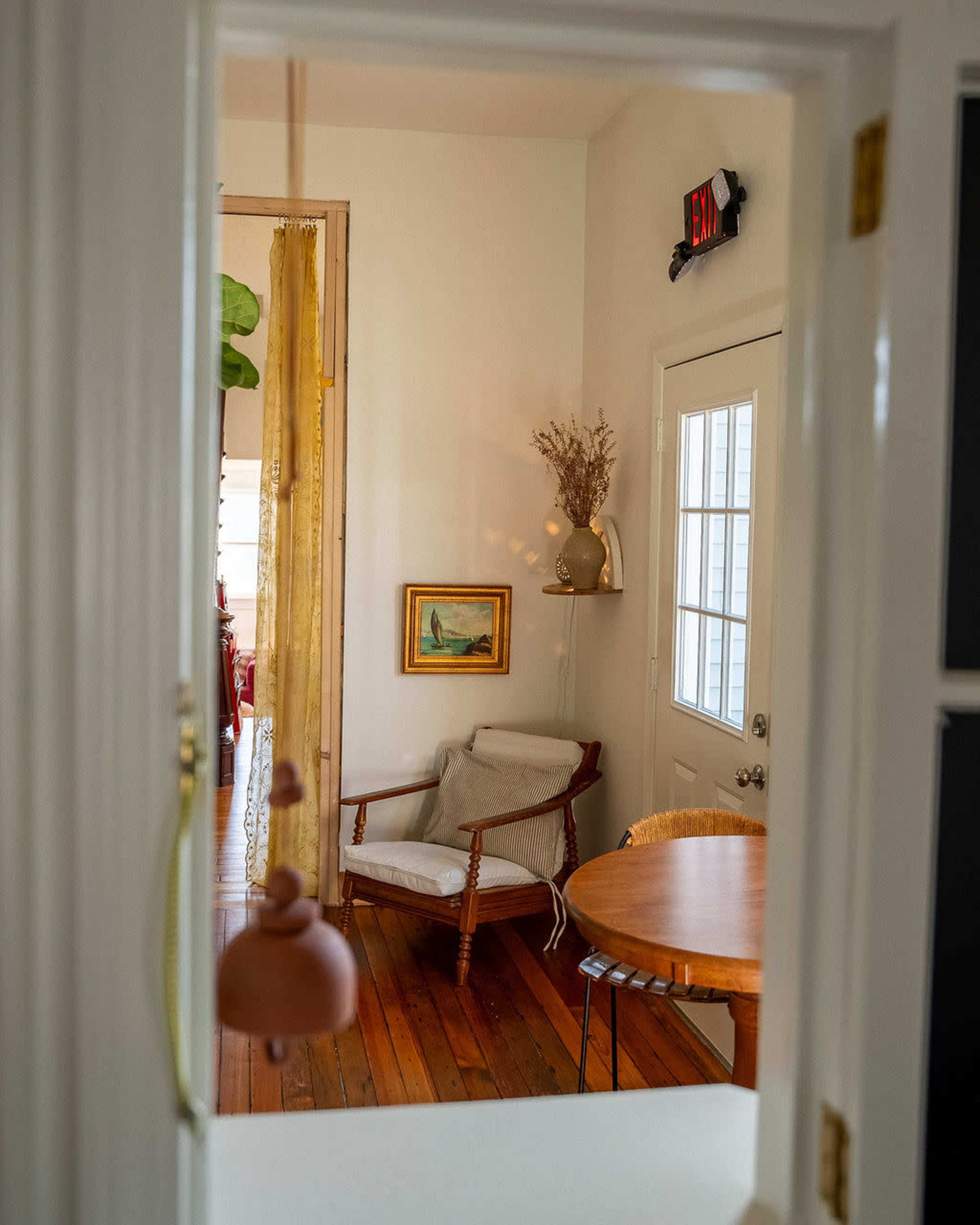 A cozy interior view shows a small room featuring a chair, a wooden table, and a door leading outside, with natural light streaming in.