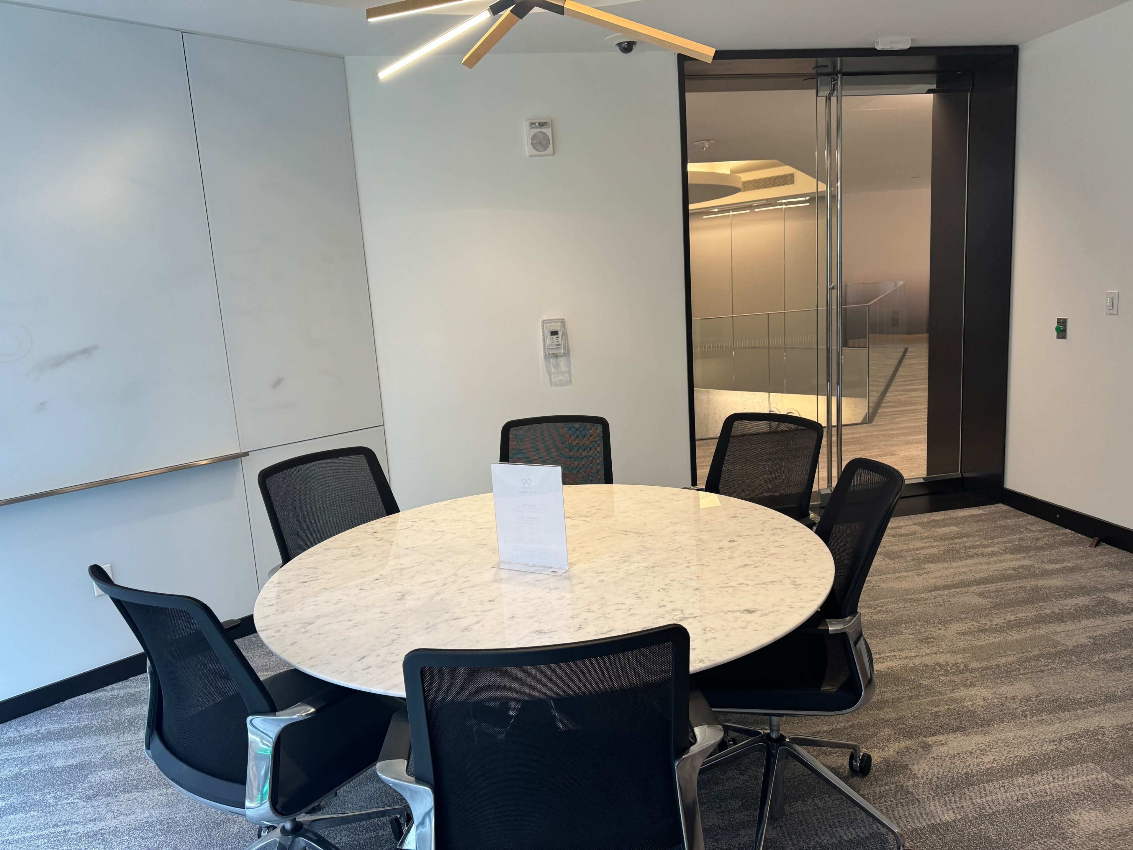 A round marble conference table surrounded by black chairs is positioned in a modern meeting room with a glass door leading to a staircase.