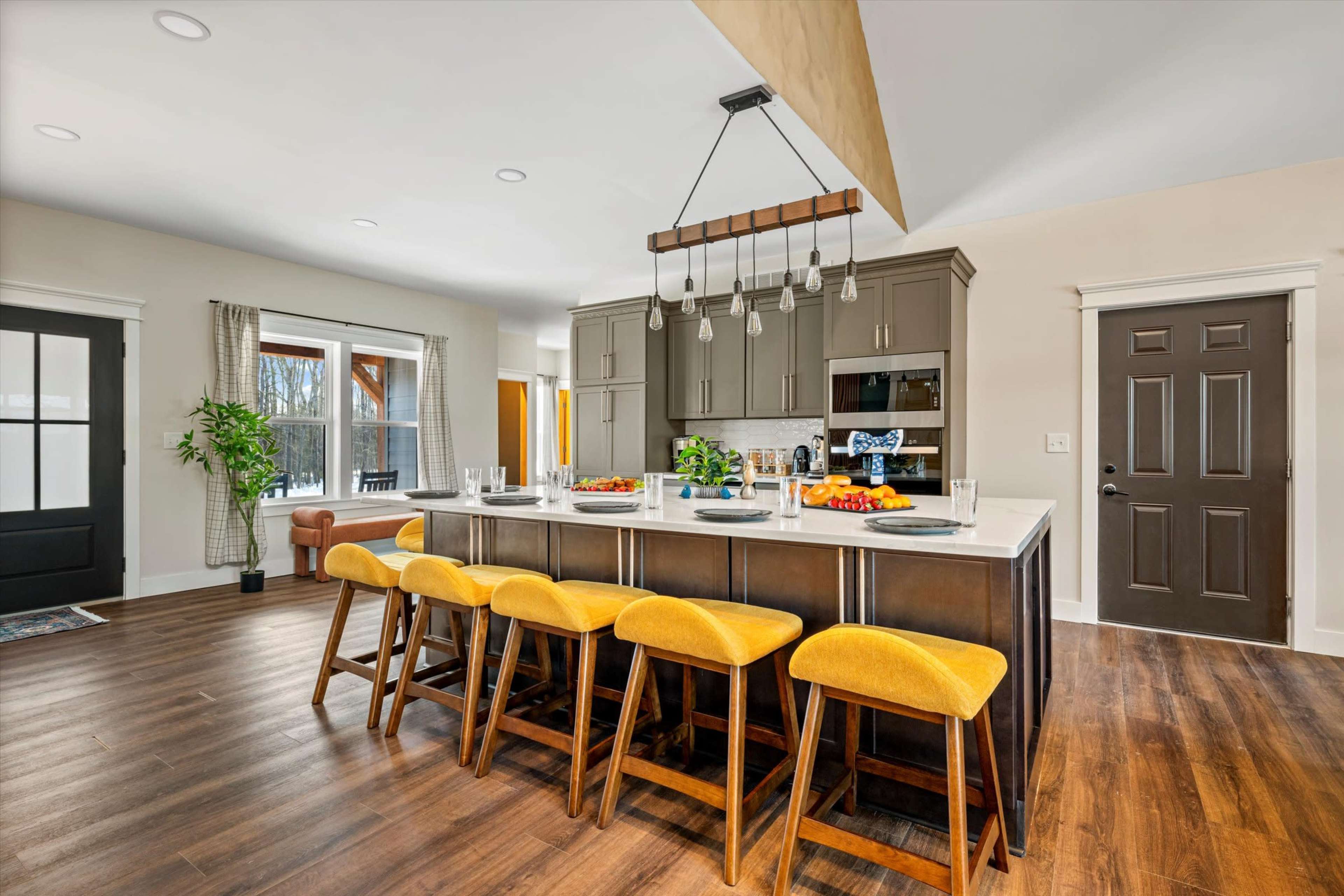 The image shows a modern kitchen with a central island and yellow bar stools, featuring dark cabinetry and large windows that provide natural light.