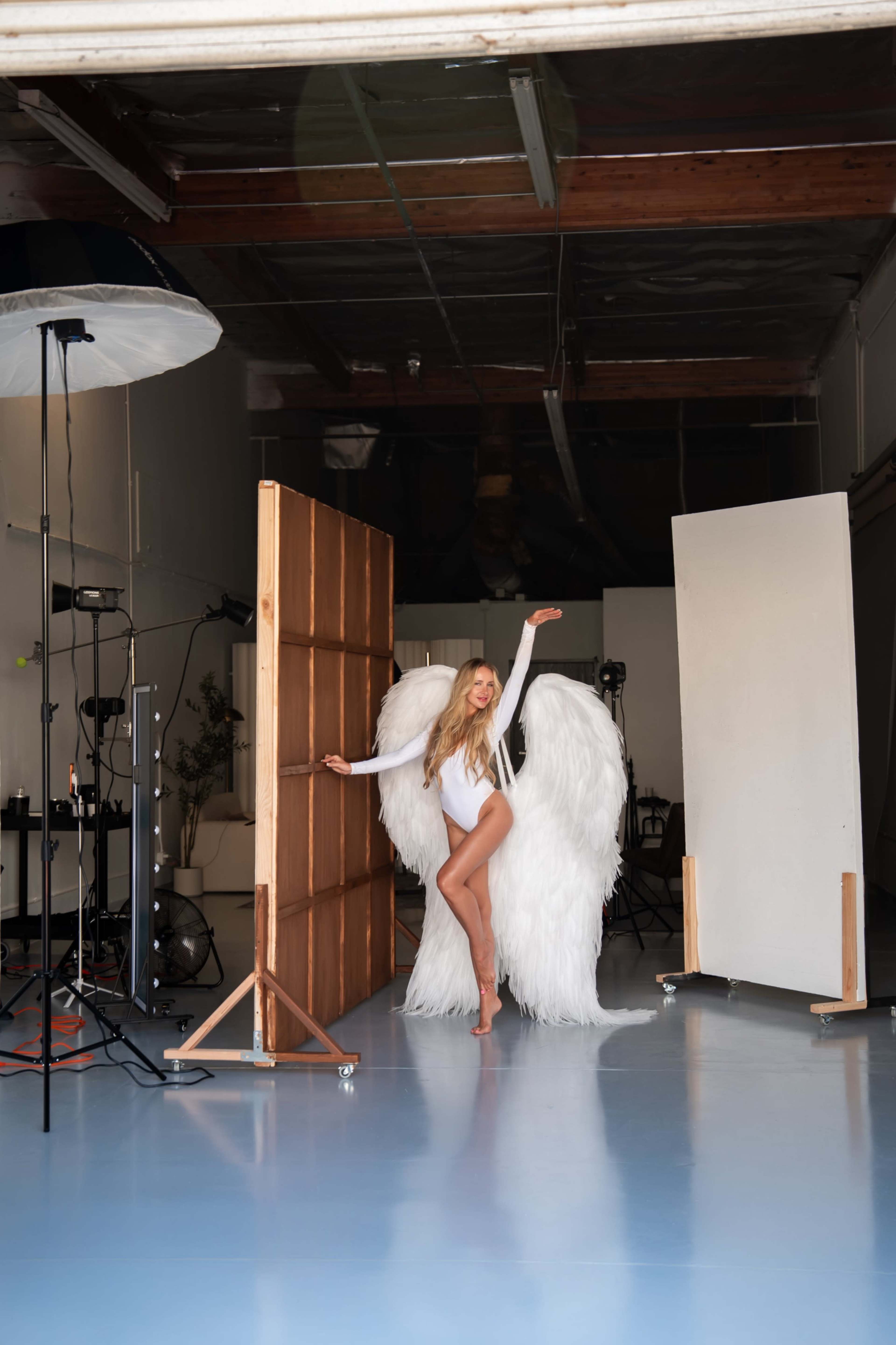 A model wearing large feathered wings poses in a studio setting with photography equipment in the background.