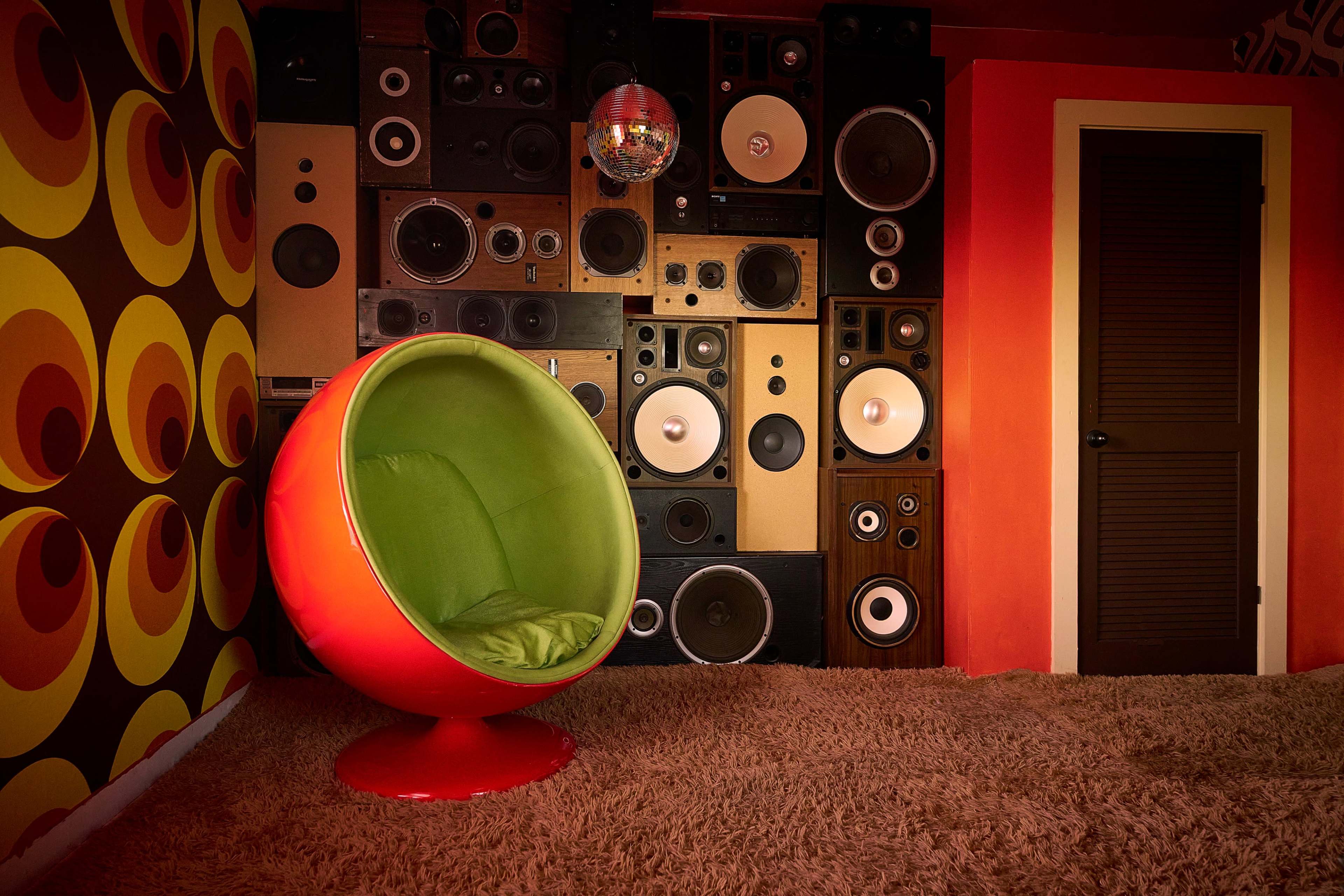 A brightly colored egg-shaped chair in front of a wall of various vintage speakers and a patterned wallpaper.