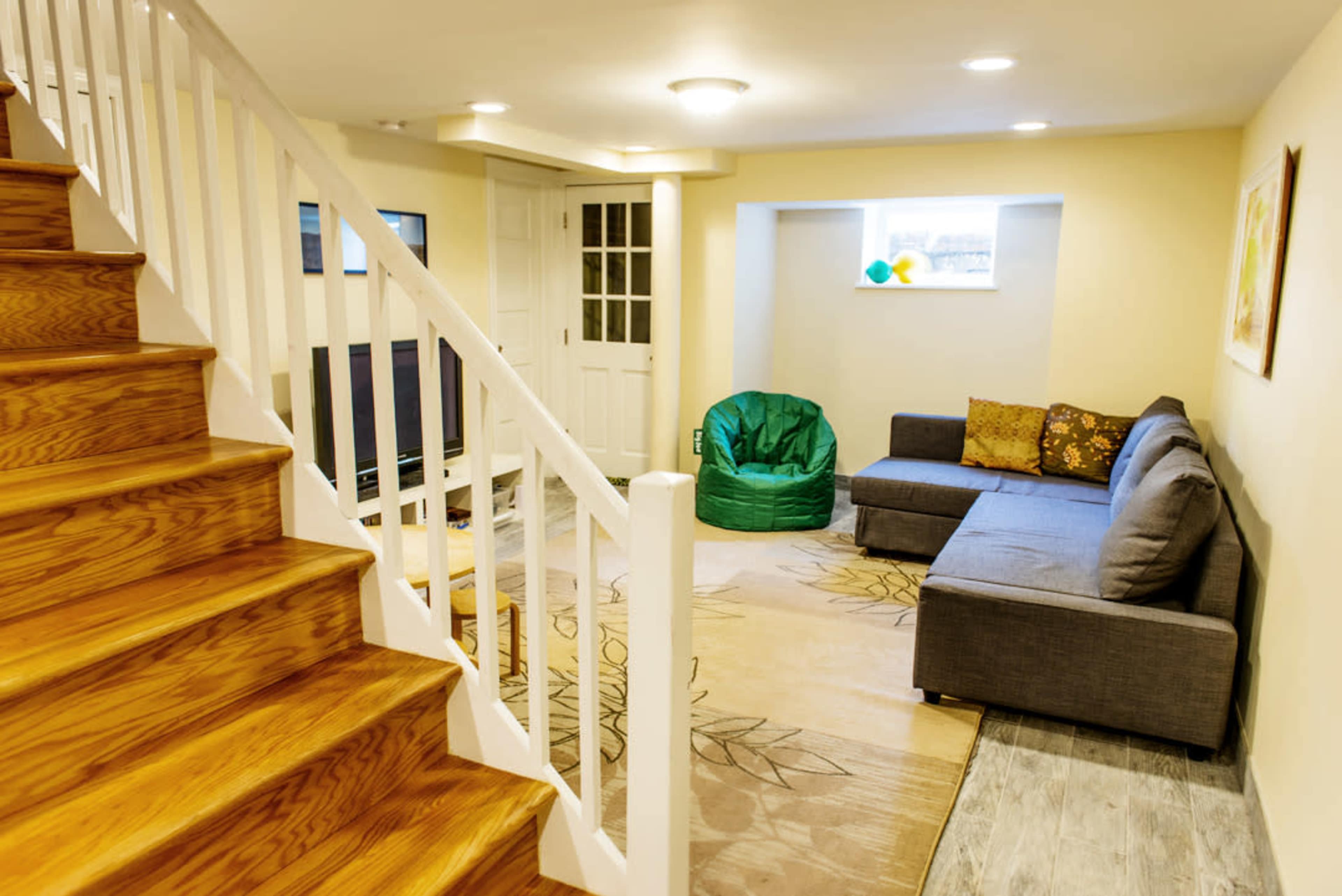 A finished basement with a staircase, a television, a green bean bag chair, and a gray sectional couch arranged on a large area rug.