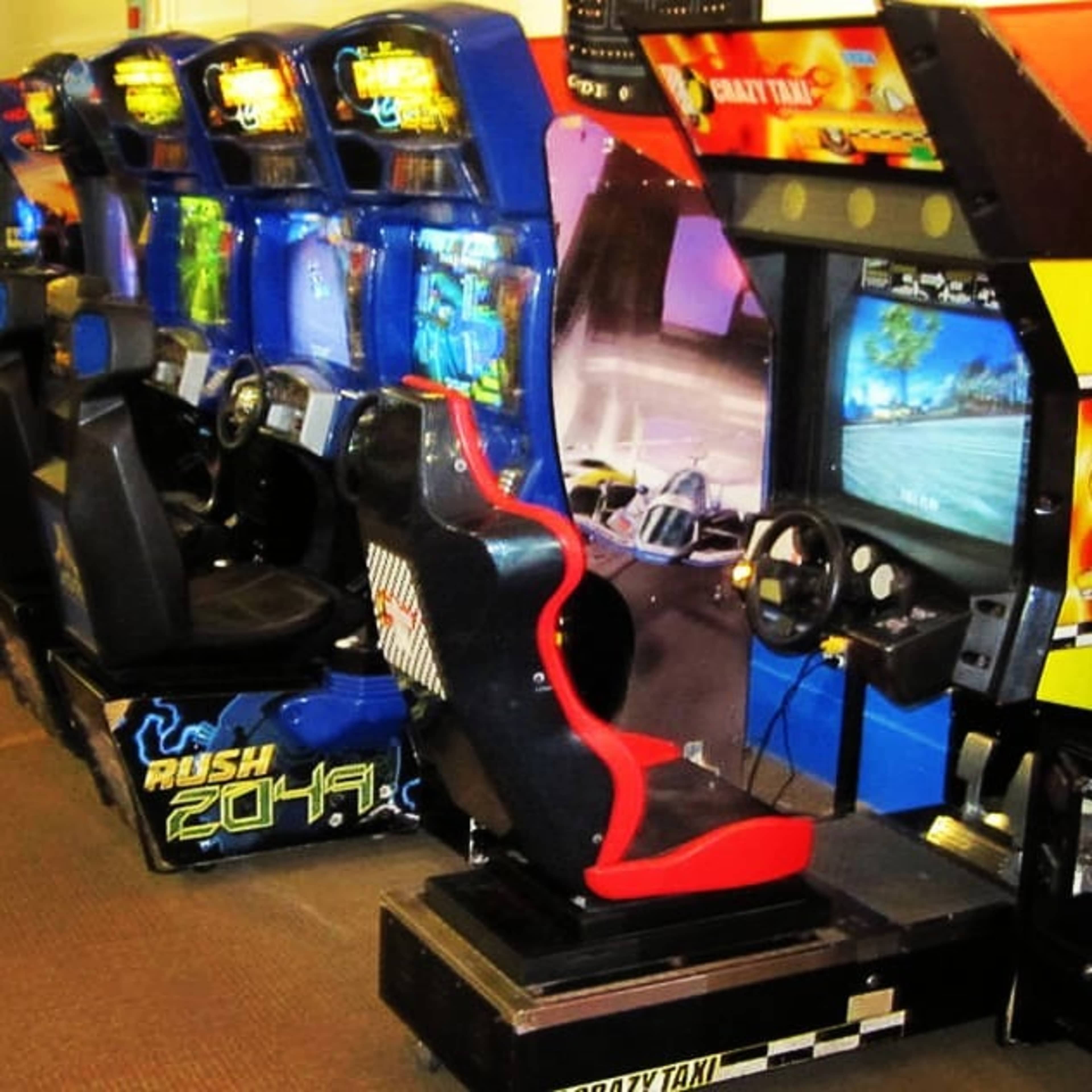 The image shows a row of arcade racing game machines, including "Rush 2049" and "Crazy Taxi," with colorful screens and driving seats.