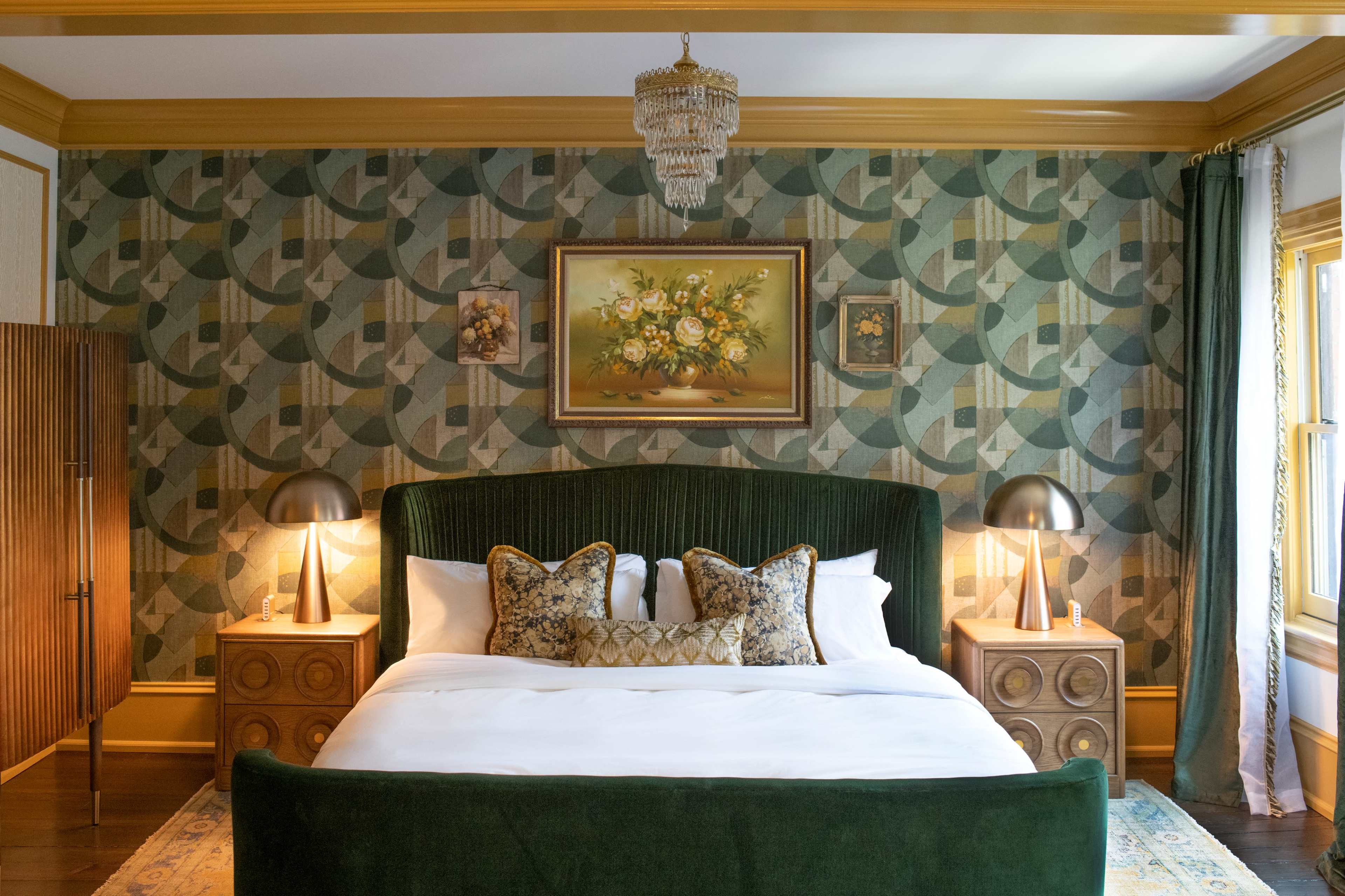 The image shows a styled bedroom featuring a large bed with an emerald green headboard, decorative pillows, two bedside lamps, and patterned wallpaper with floral artwork.
