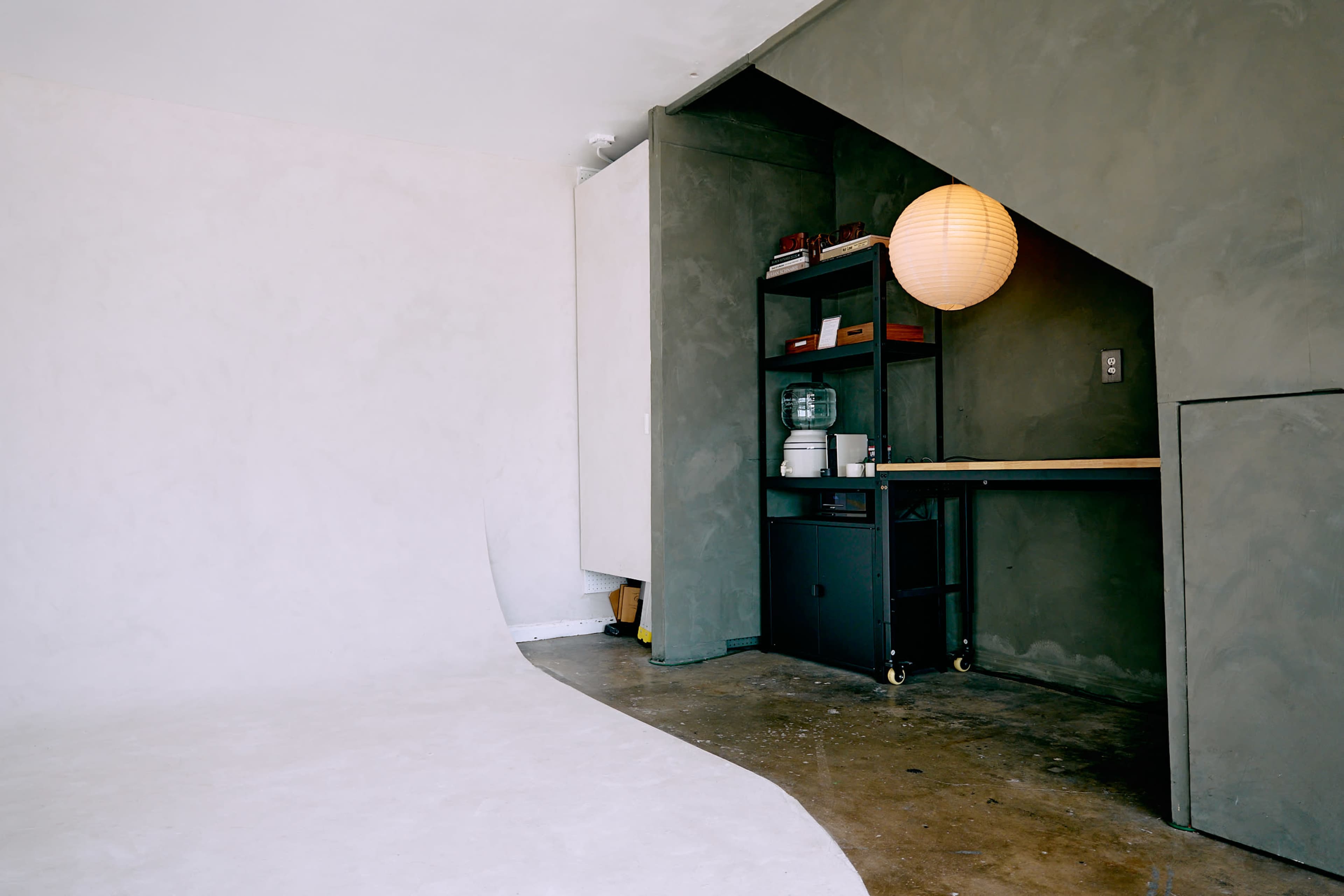 The image shows a minimalist interior with a curved white backdrop, a concrete wall, and a shelving unit under a staircase featuring a round paper lantern.