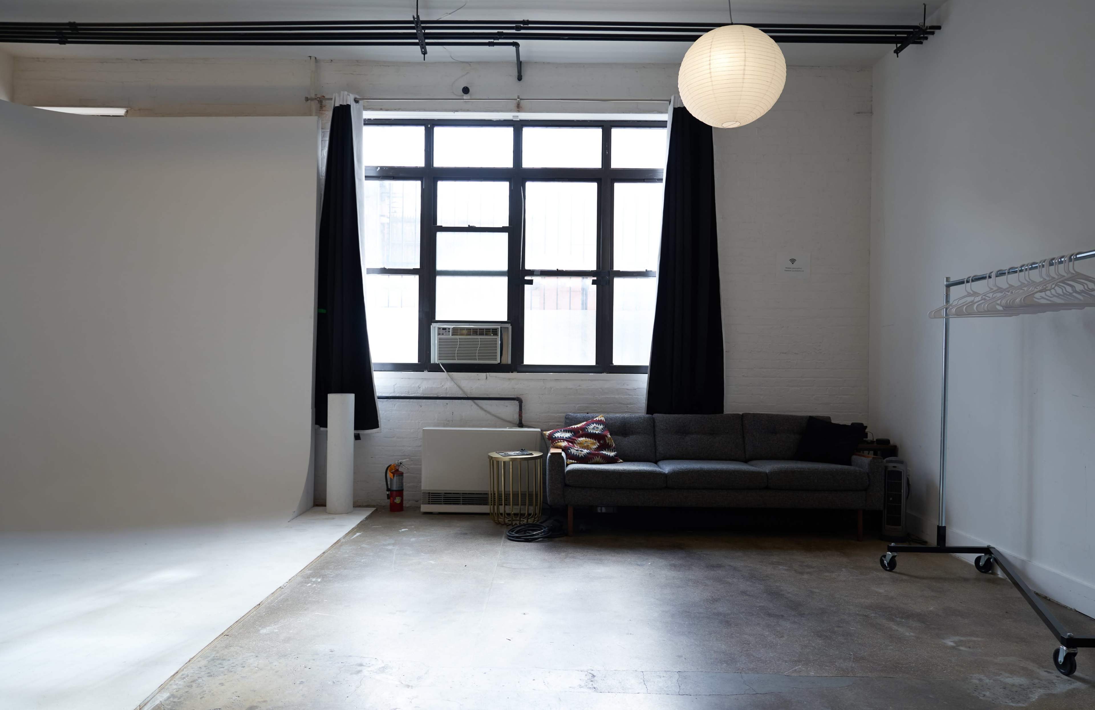 The image shows an empty studio space with a gray sofa, a large window with black curtains, and a round paper lamp hanging from the ceiling.