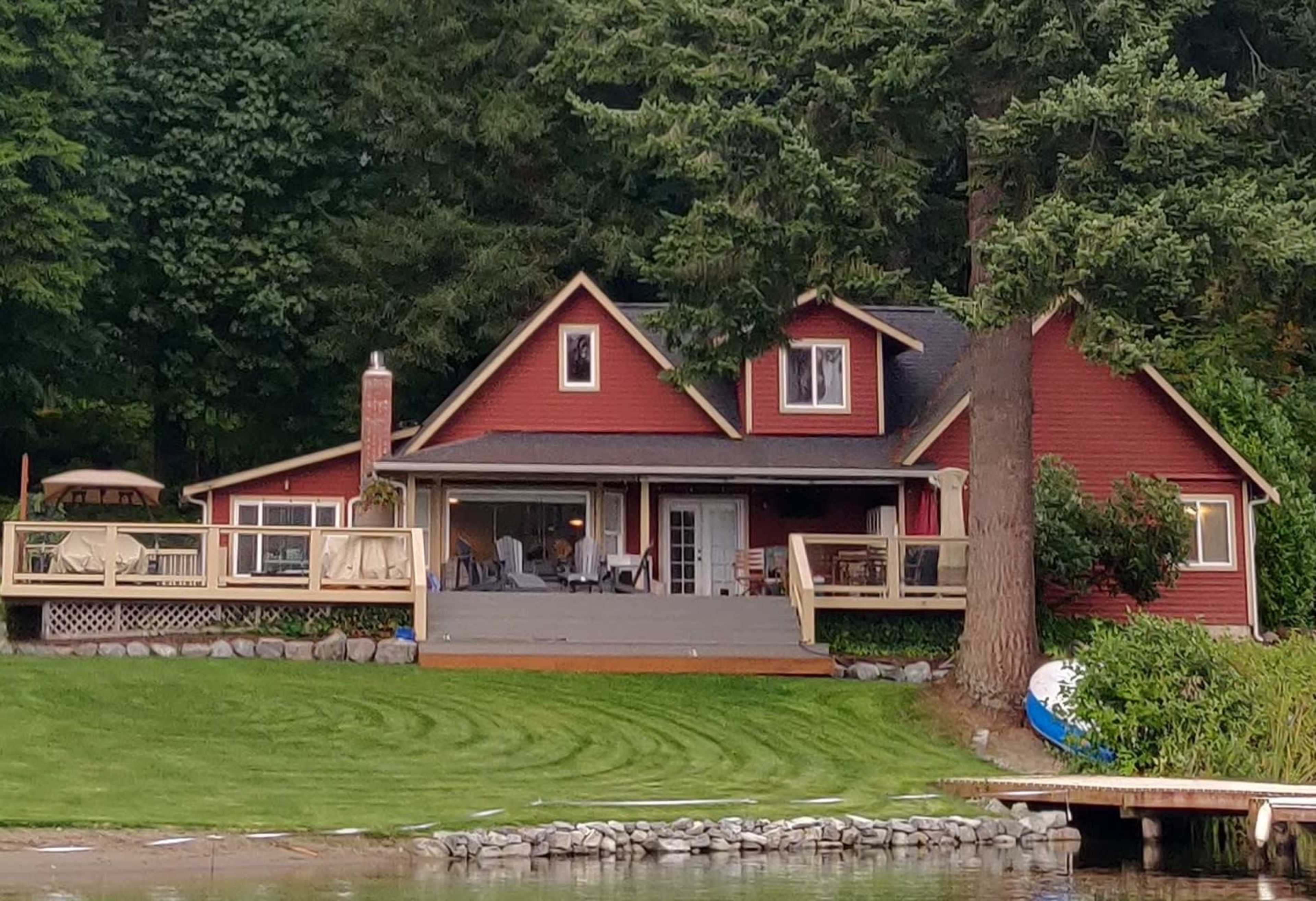 A red cottage with a porch and a large lawn sits by a body of water, surrounded by trees.