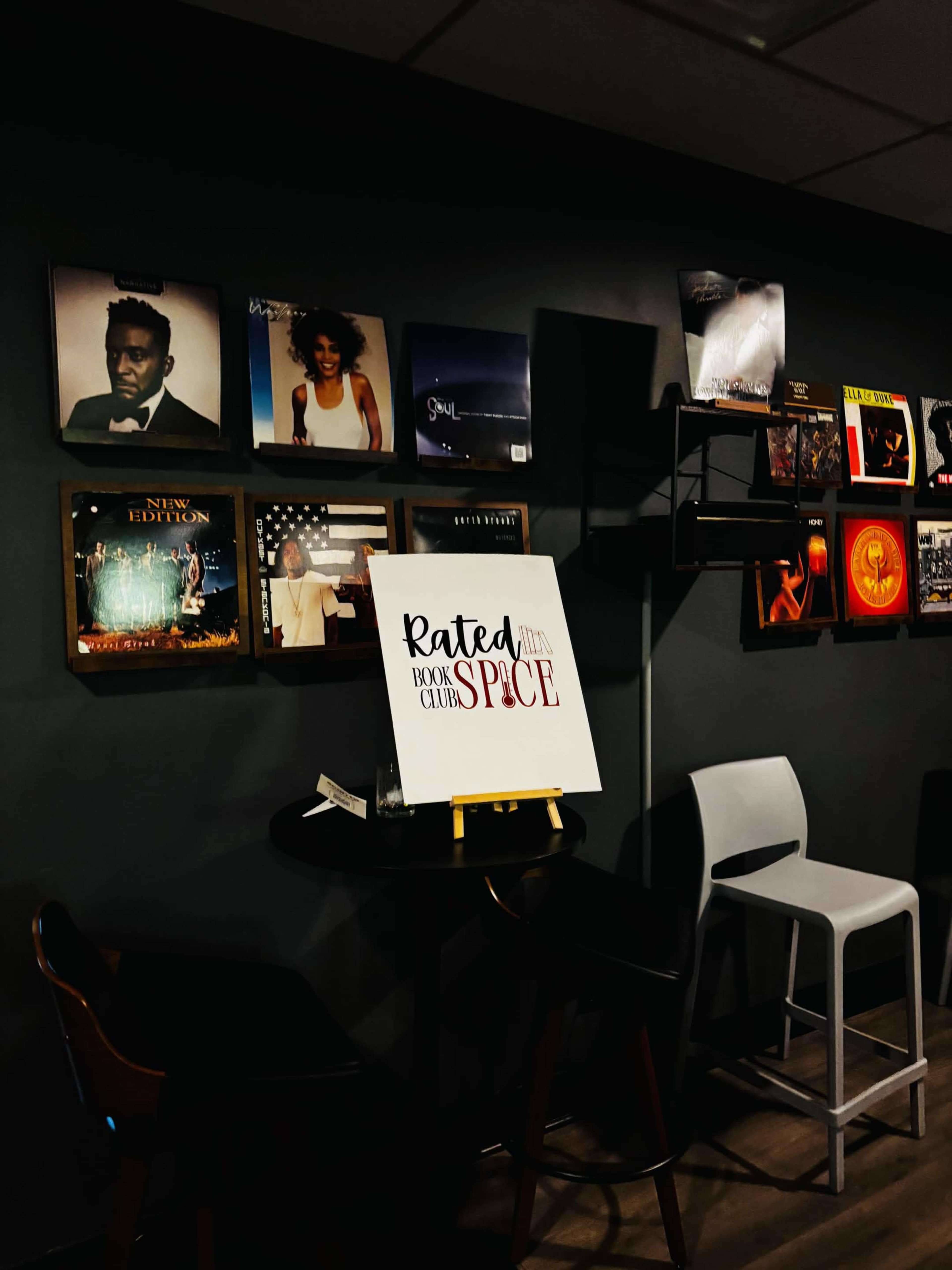 A small table with a sign reading "Rated Book Club" is positioned in front of a wall adorned with framed vinyl records and shelves displaying various music albums.