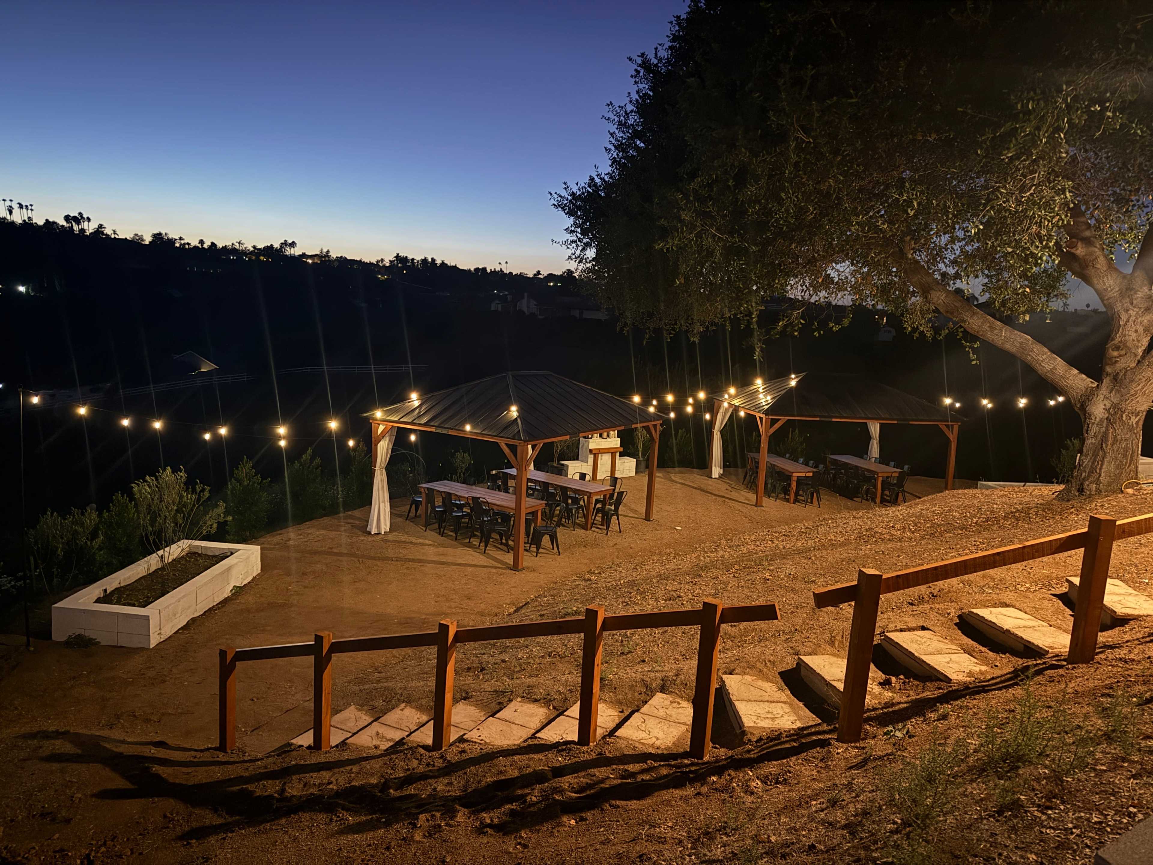 The image shows a hillside outdoor patio area with string lights overhead, featuring several tables and a peaceful sunset in the background.