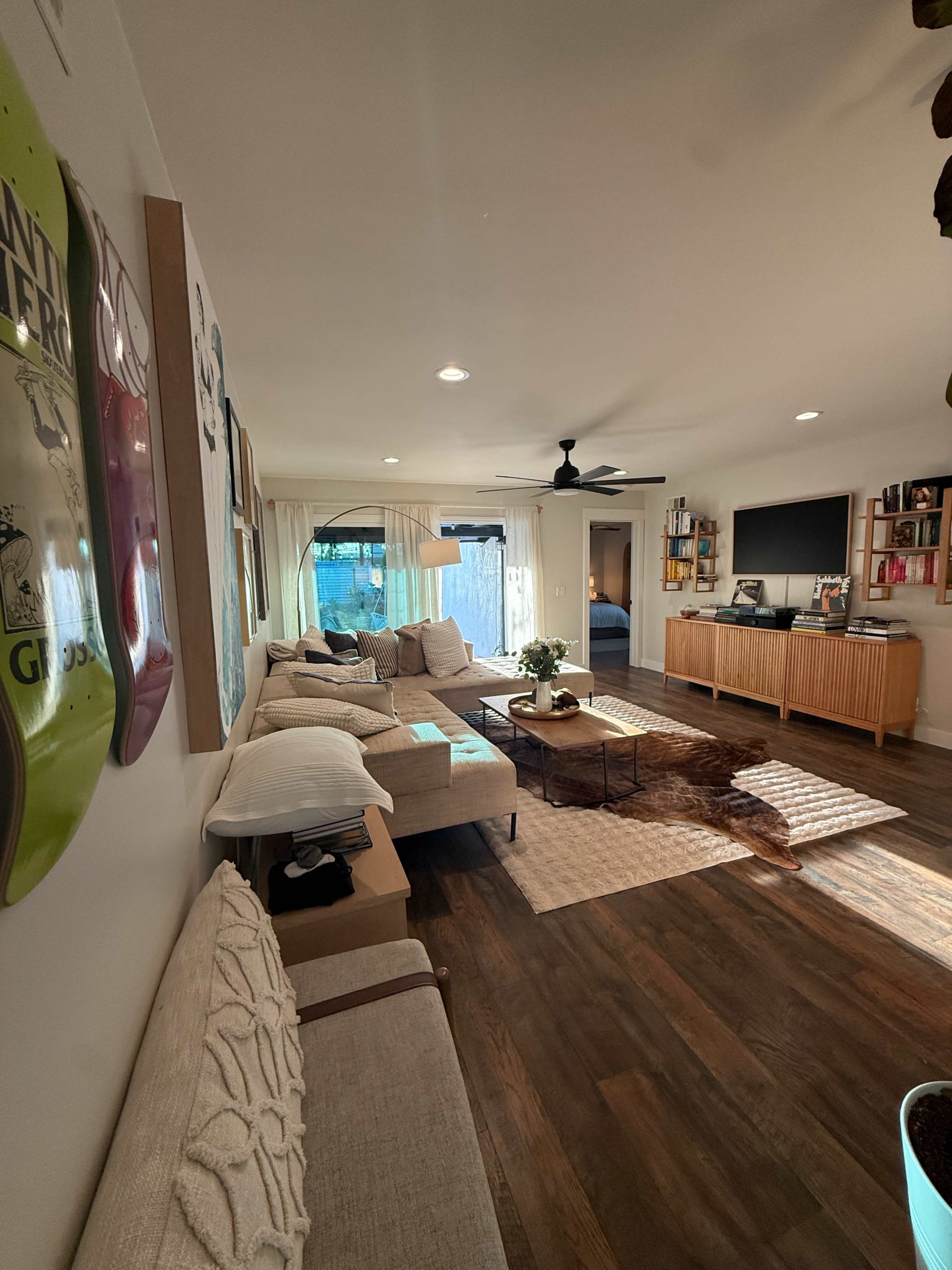 The image shows a well-lit living room featuring a sectional sofa, a coffee table on a light rug, and a television mounted on the wall opposite a wooden cabinet.