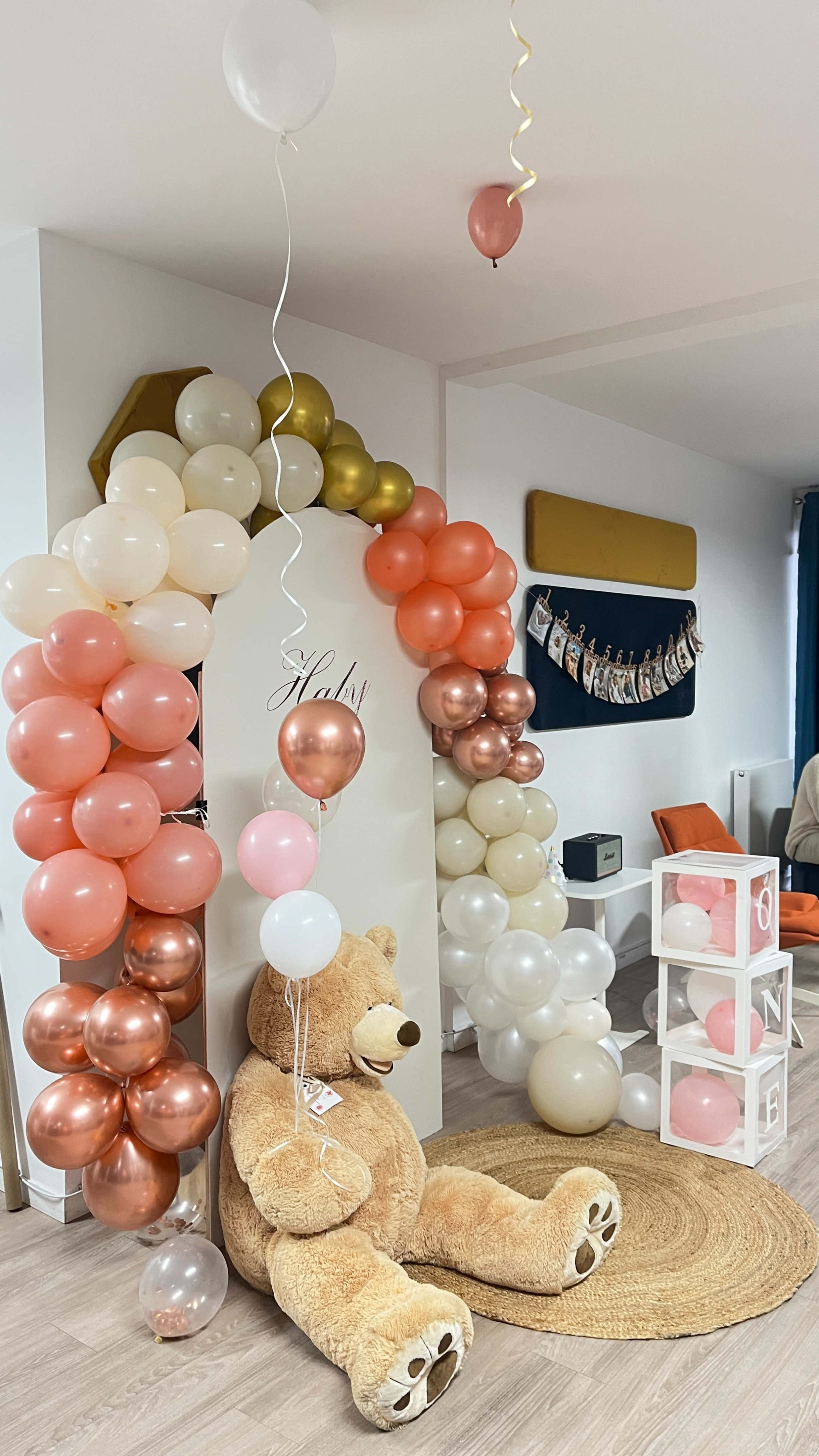 A large teddy bear sits on a rug under a decorative arch made of balloons in various colors, with a wall displaying photographs in the background.