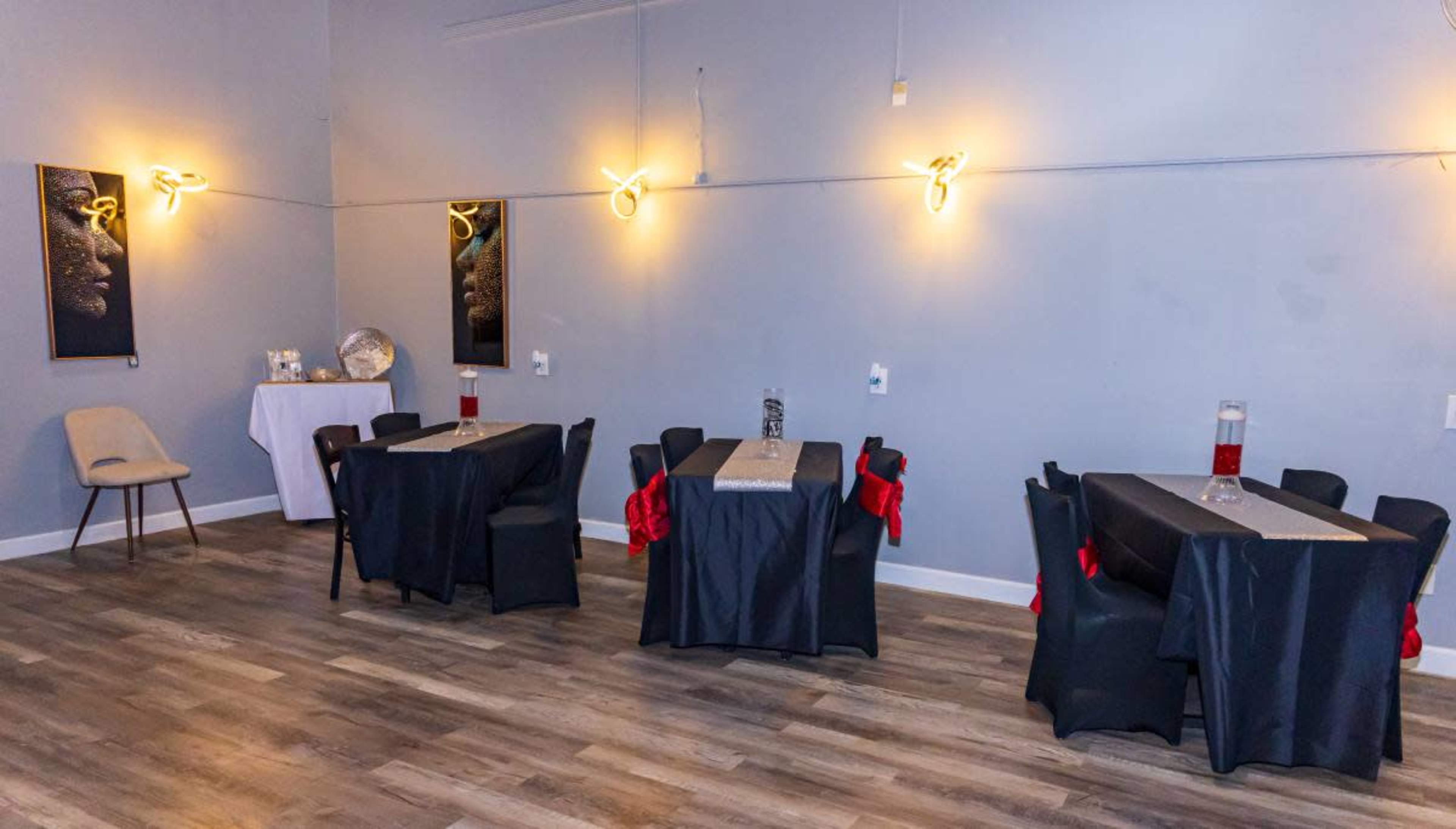 The image shows a spacious, minimally decorated room with several black tables covered in dark tablecloths, each set with a centerpiece and red accents, and illuminated by modern light fixtures.