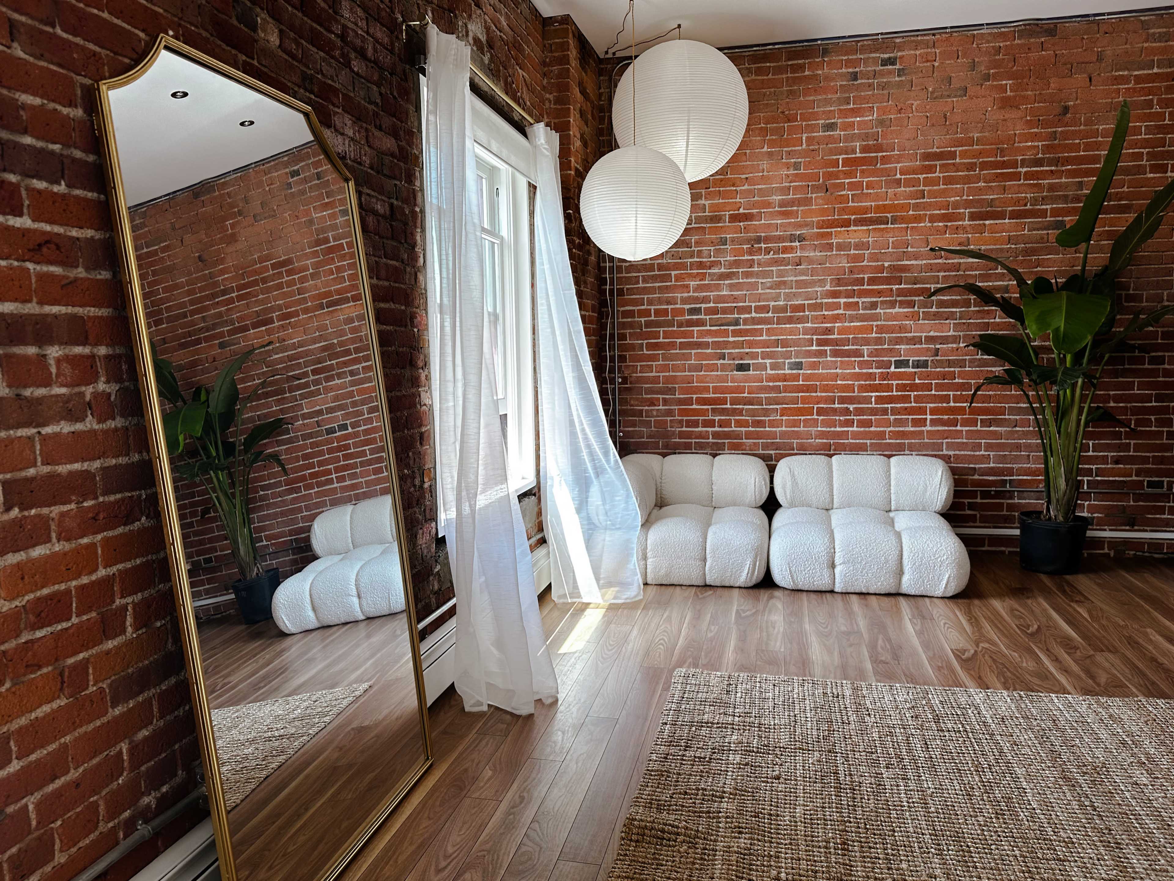 The image shows a room with exposed brick walls, a large mirror, a white seating area, and two lantern-style light fixtures hanging from the ceiling.