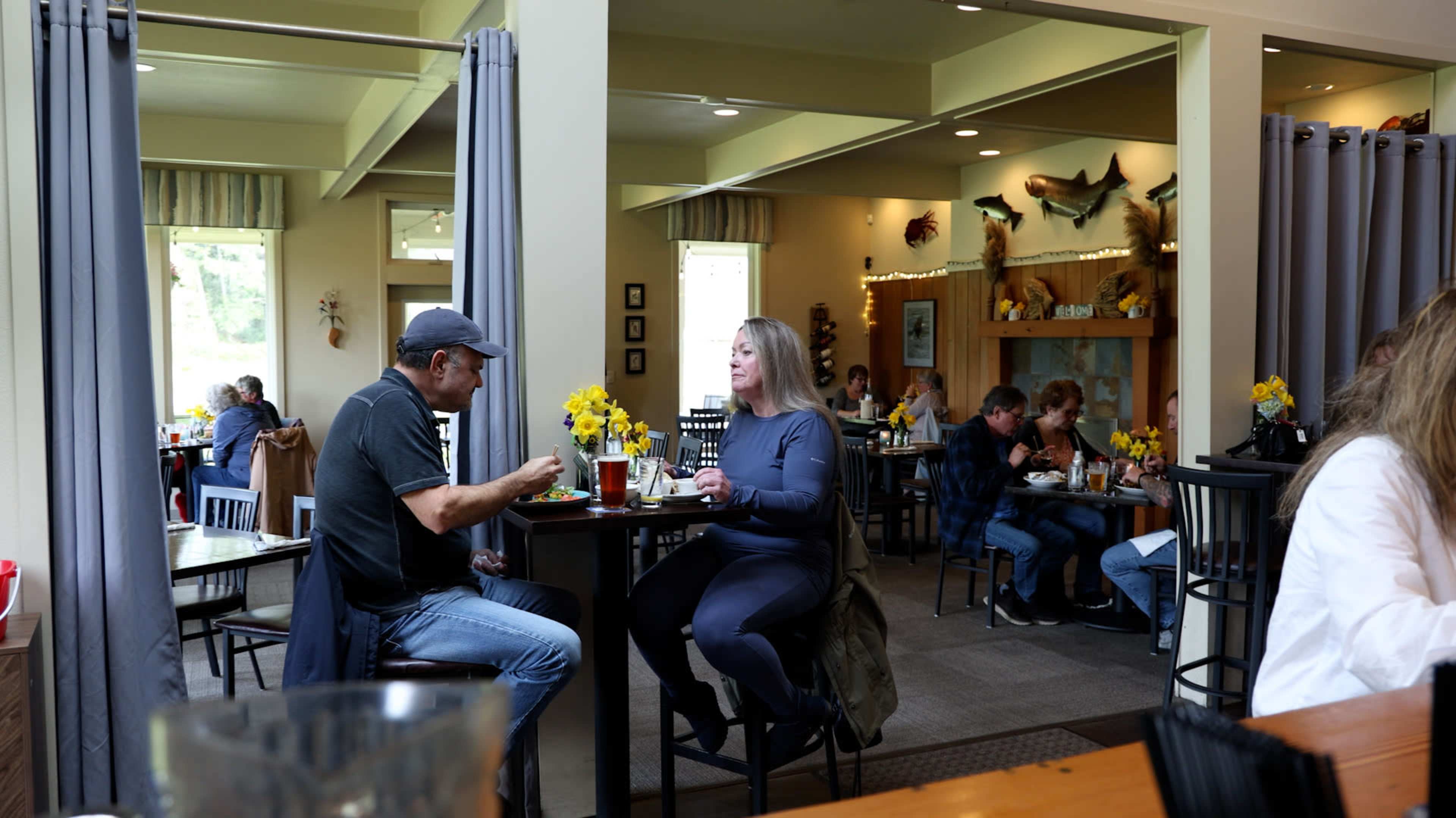 Two diners engage in conversation at a small table in a busy restaurant, while other patrons enjoy their meals in the background.