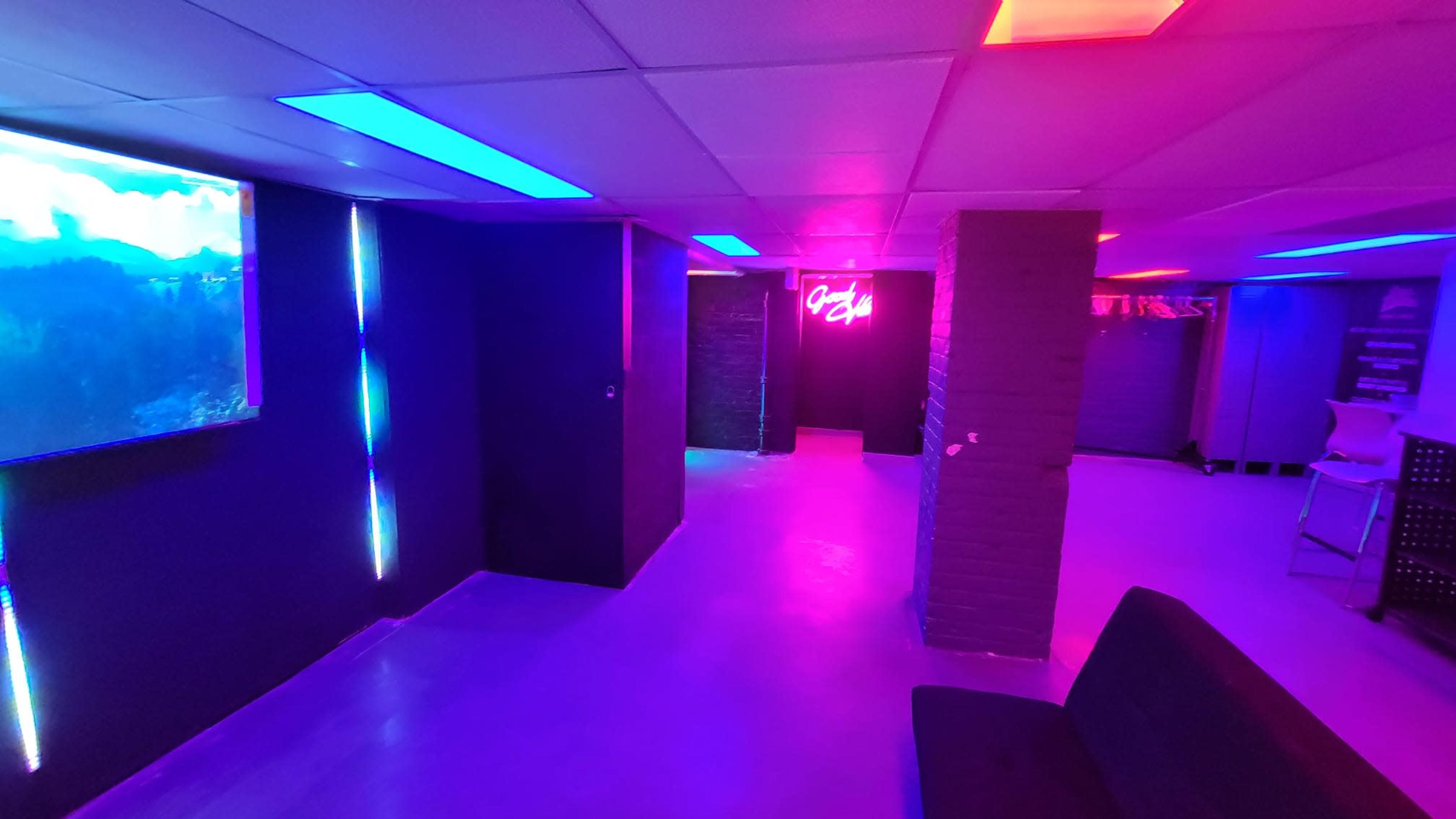 The scene features a dimly lit room with colorful LED lighting, including neon blue and pink accents, and a large screen displaying a scenic image on one wall.