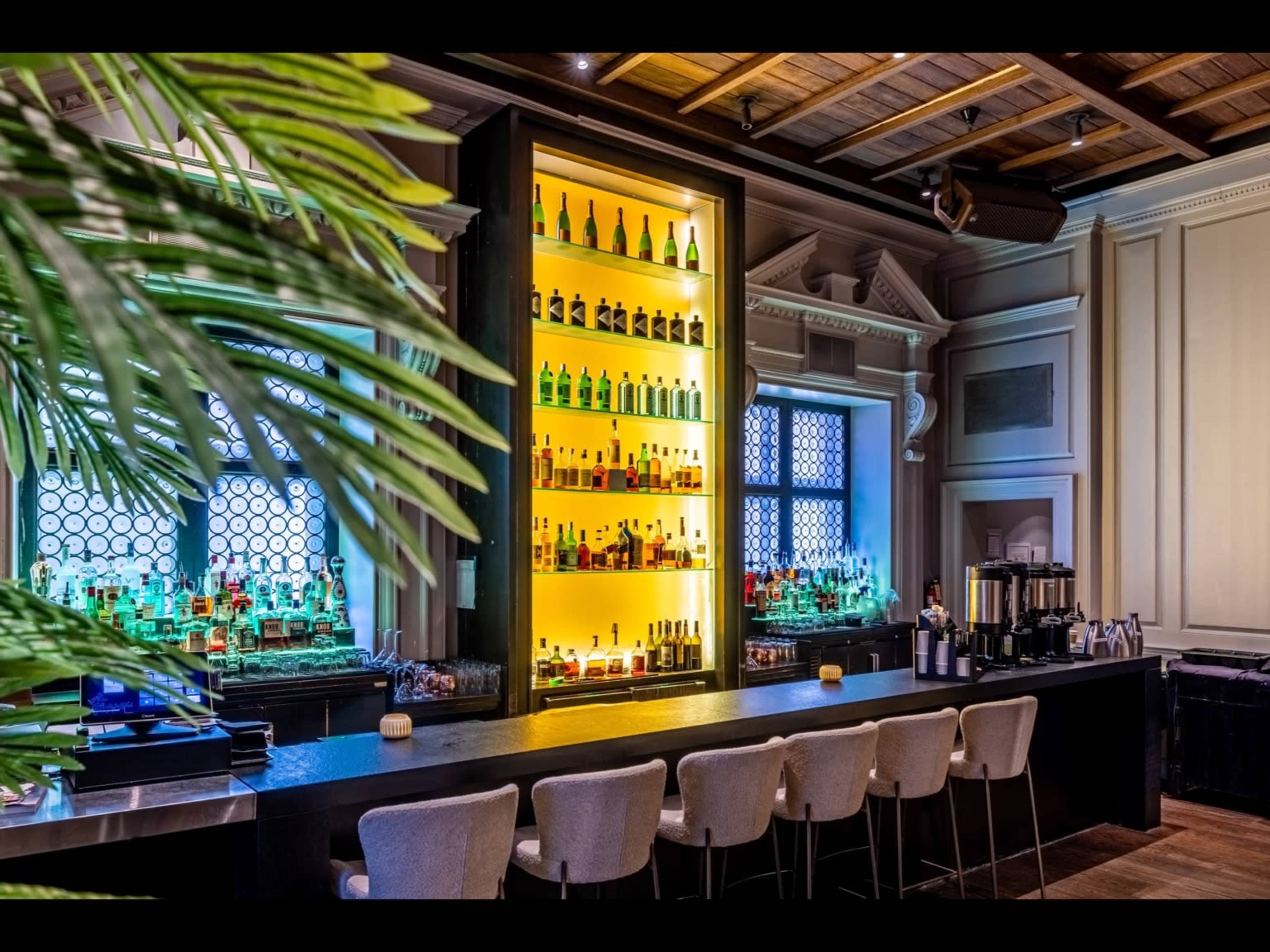 The image shows a modern bar with a well-lit shelf displaying various bottles of alcohol against a backdrop of large windows.