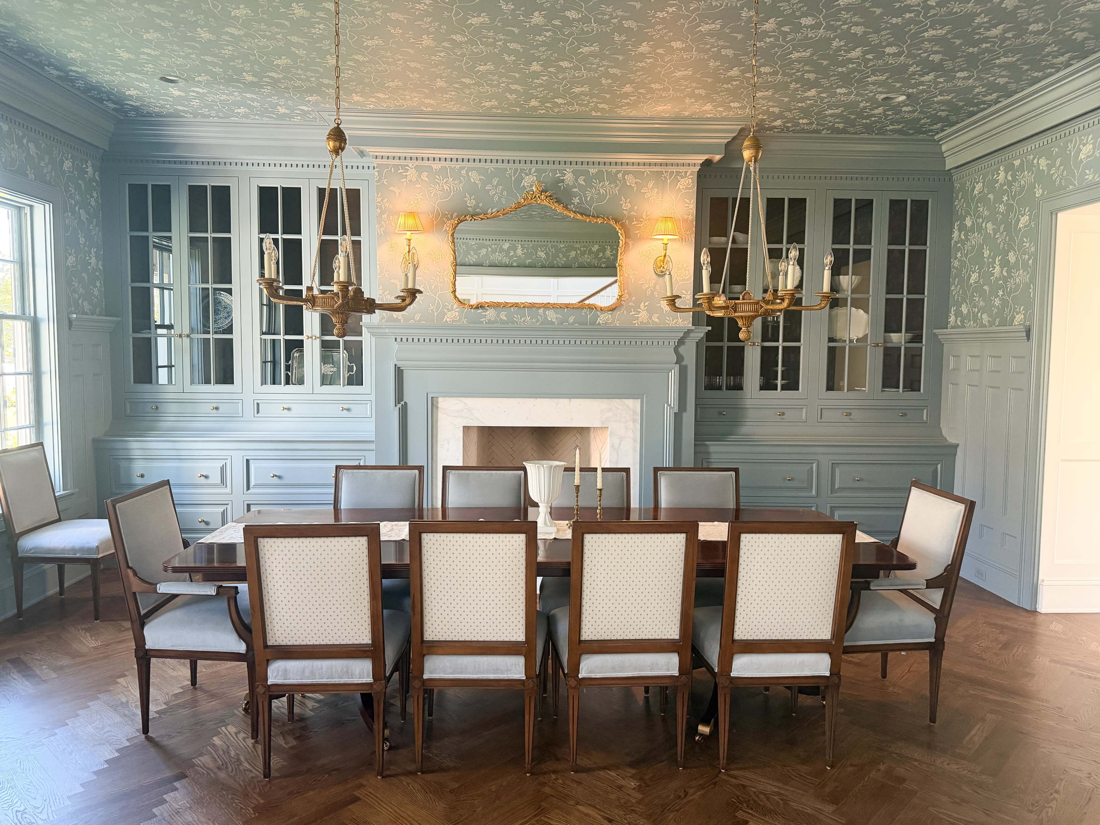 The image shows a spacious dining room featuring a long wooden table surrounded by twelve chairs, with elegant cabinetry and a fireplace decorated by soft-colored walls and vintage light fixtures.
