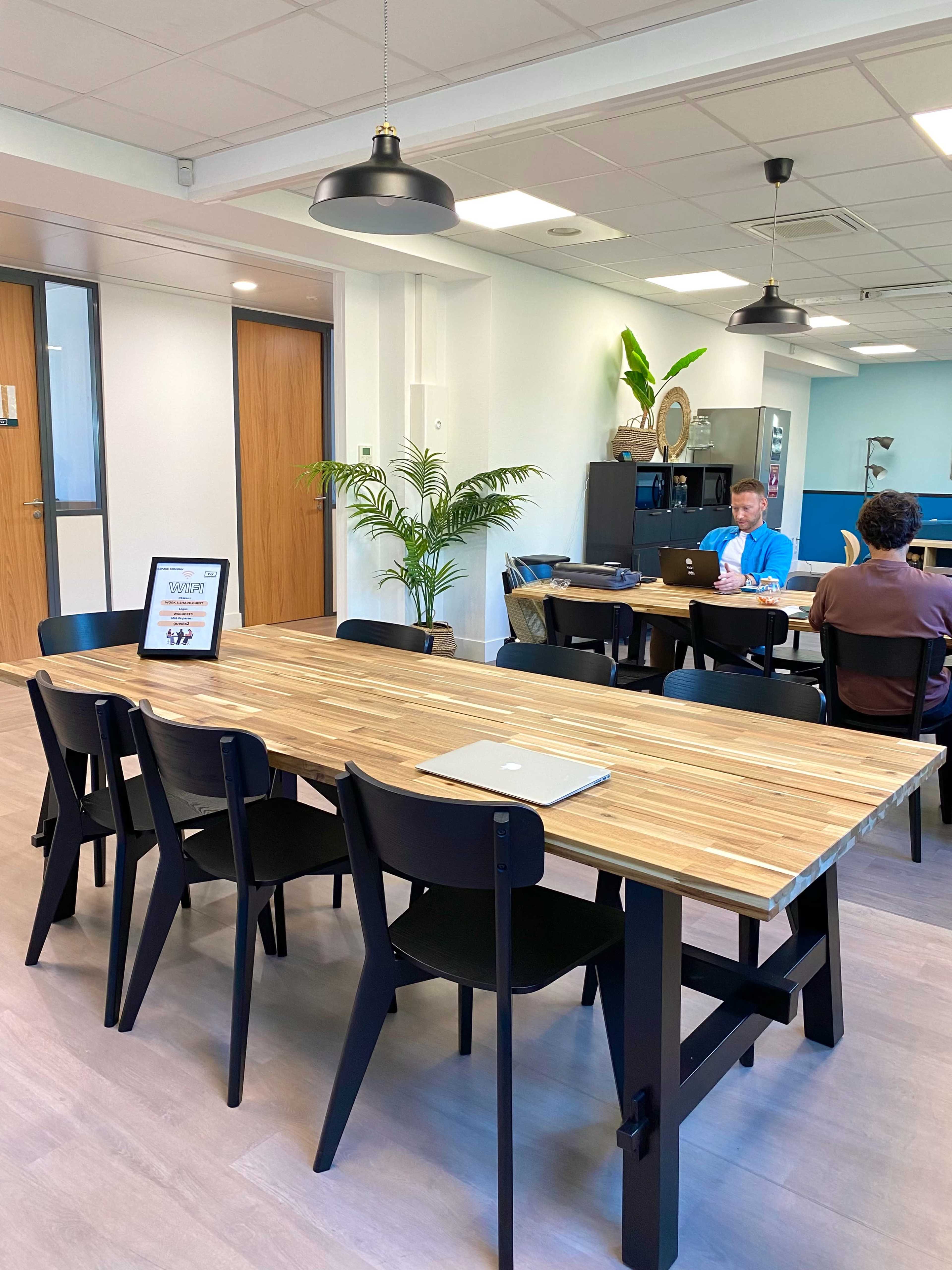 A modern office space with a large wooden table surrounded by black chairs, where two people are working on laptops.