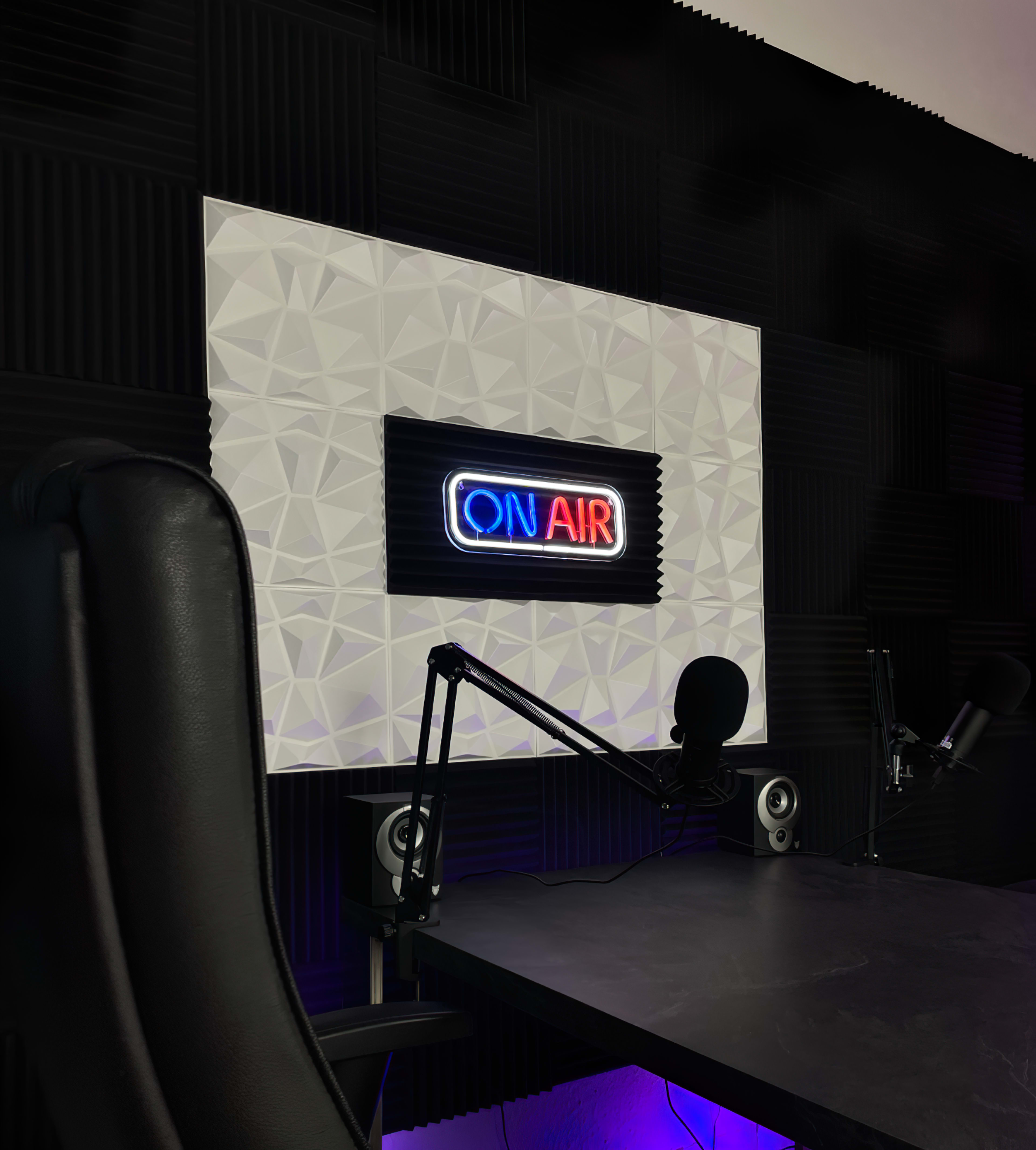 A radio studio setup featuring a "ON AIR" neon sign, a black desk with microphones, and soundproof wall panels.