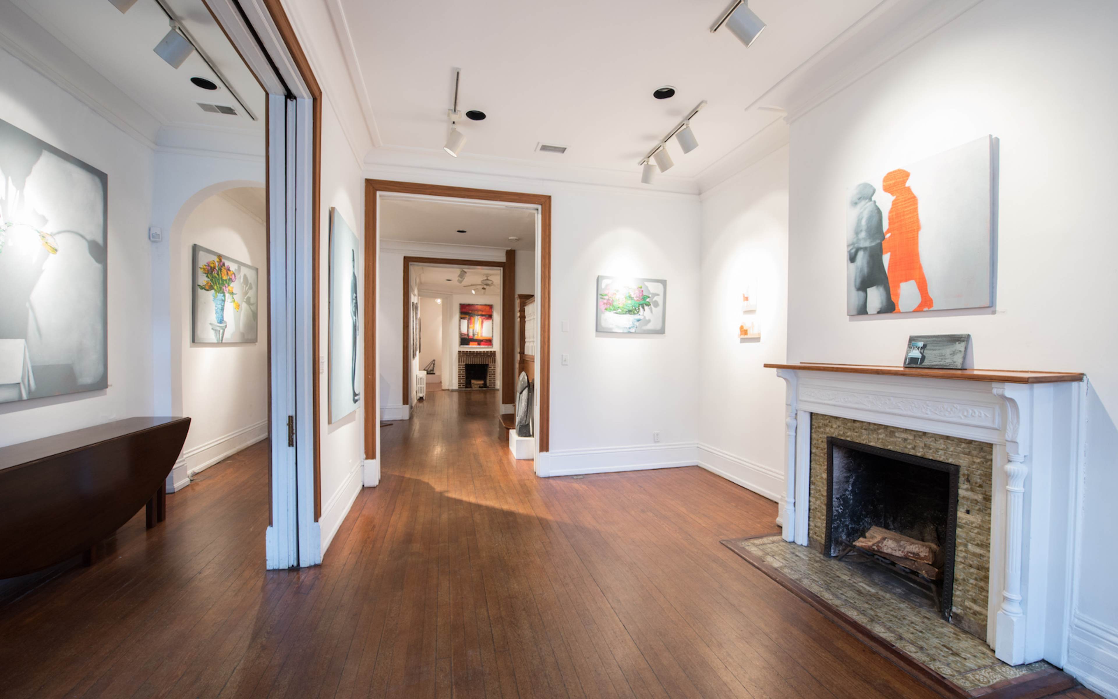 A well-lit gallery features wooden floors and white walls adorned with various artworks, including a fireplace at the center.