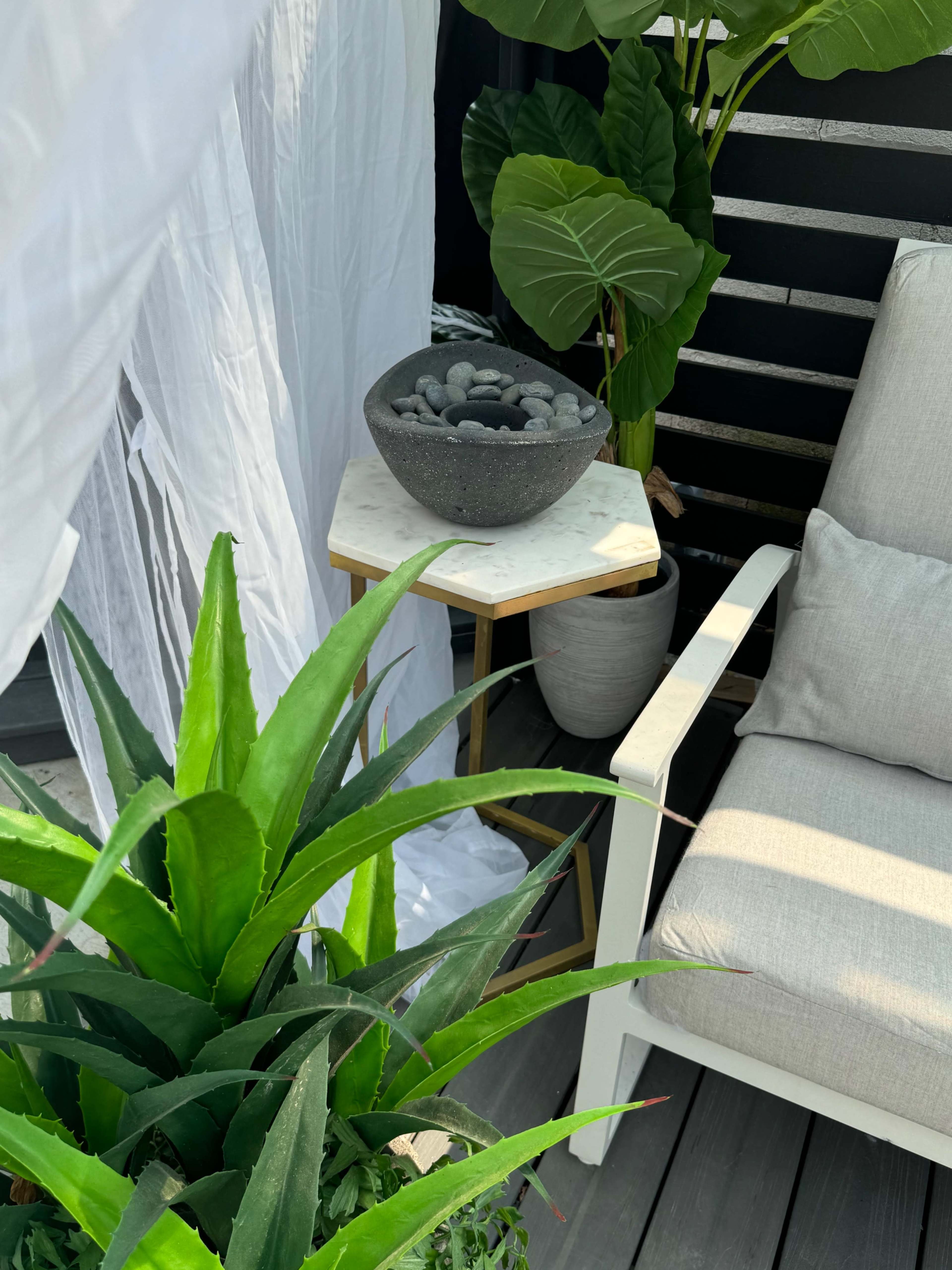 A white curtain drapes beside a gray patio chair and a small table with a decorative bowl, surrounded by large green plants.