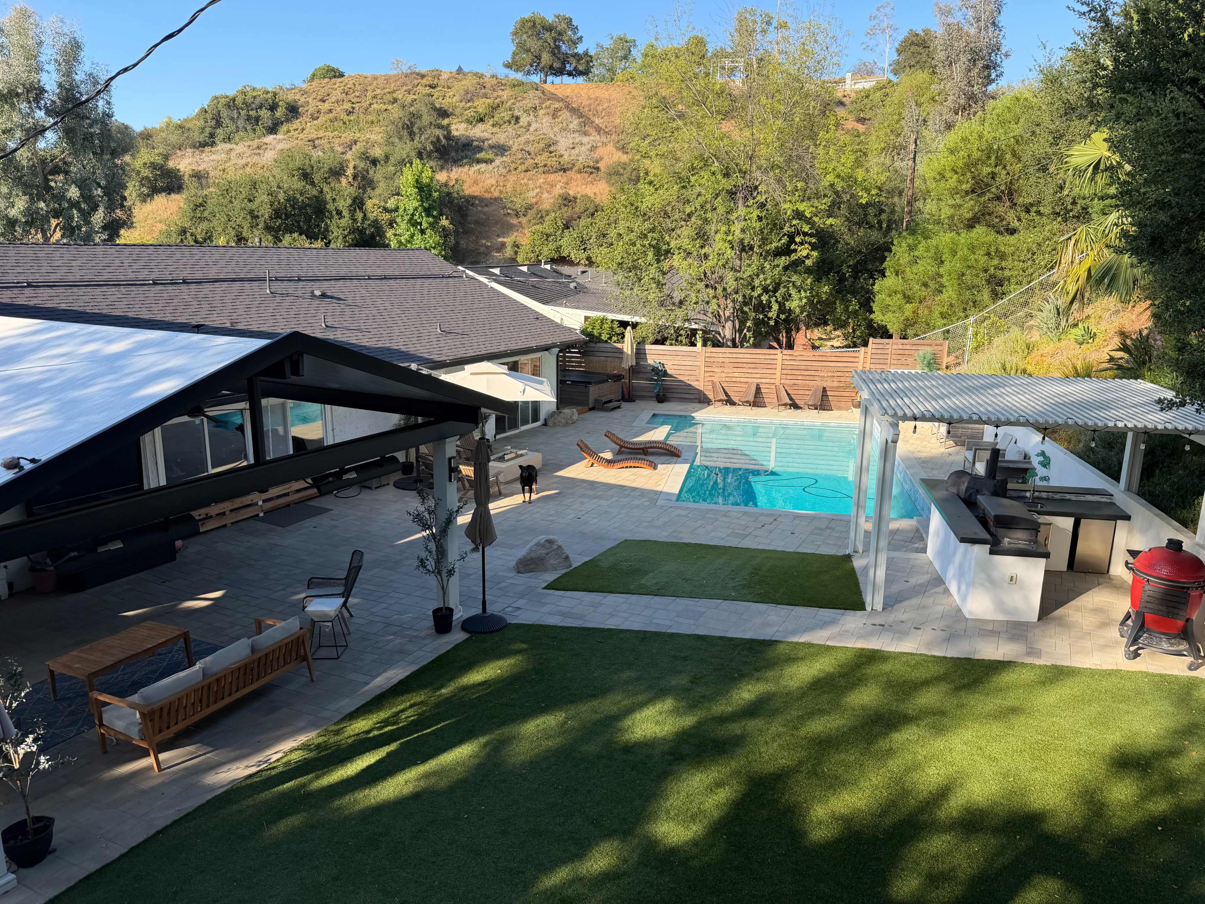 The image shows a modern backyard featuring a swimming pool, outdoor kitchen, and seating area surrounded by greenery and a hillside.