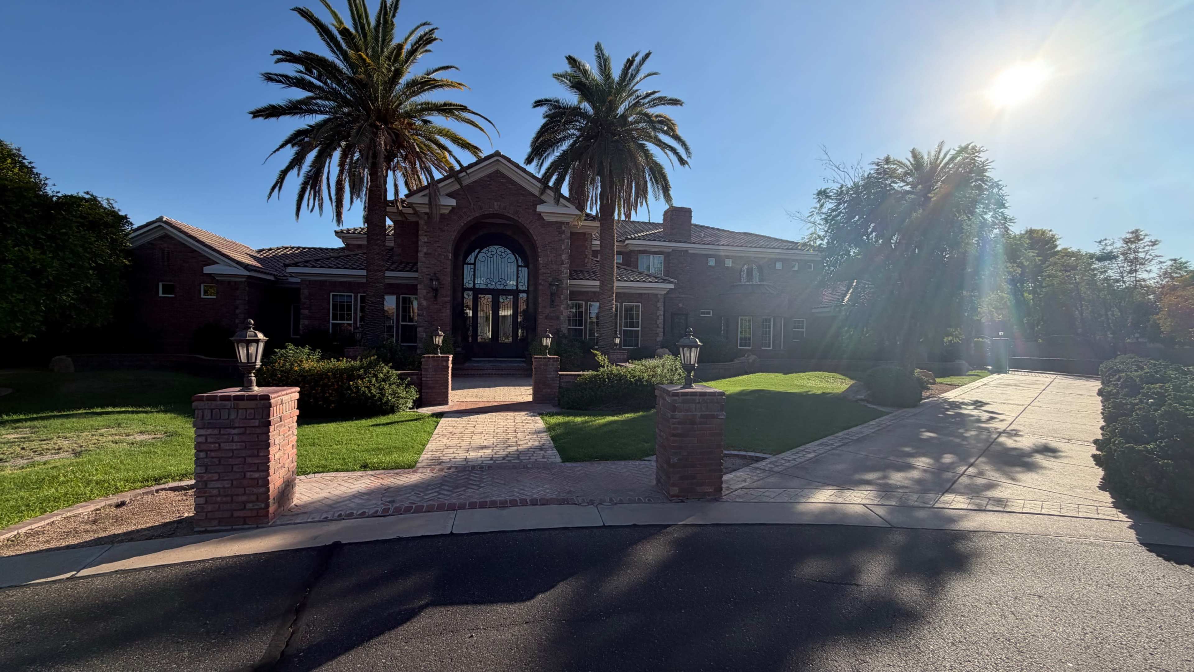 A grand brick mansion with palm trees in front, featuring a large entrance and a well-maintained lawn.