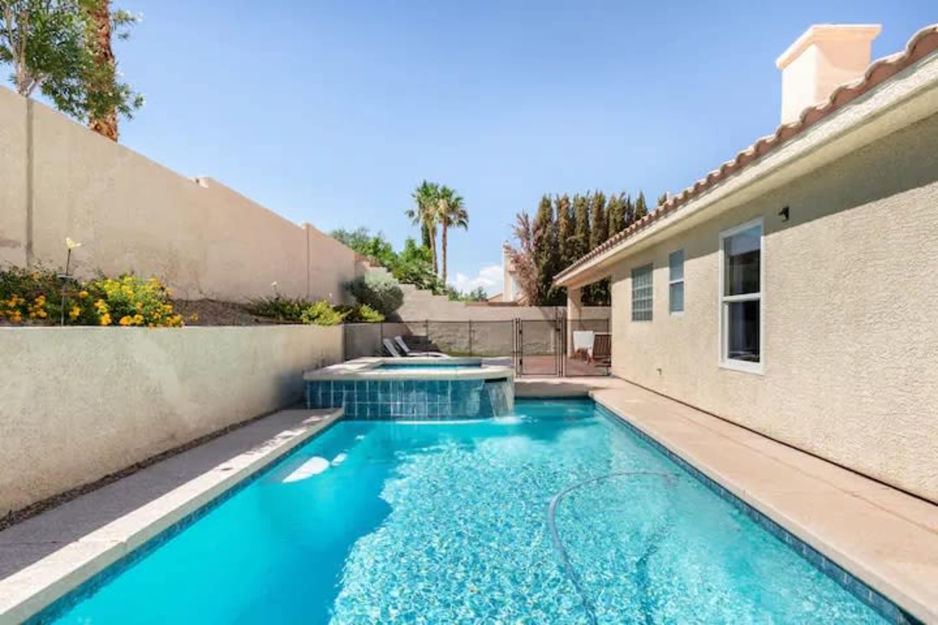 A swimming pool with a spa, bordered by a wall and gardens, adjacent to a single-story home.