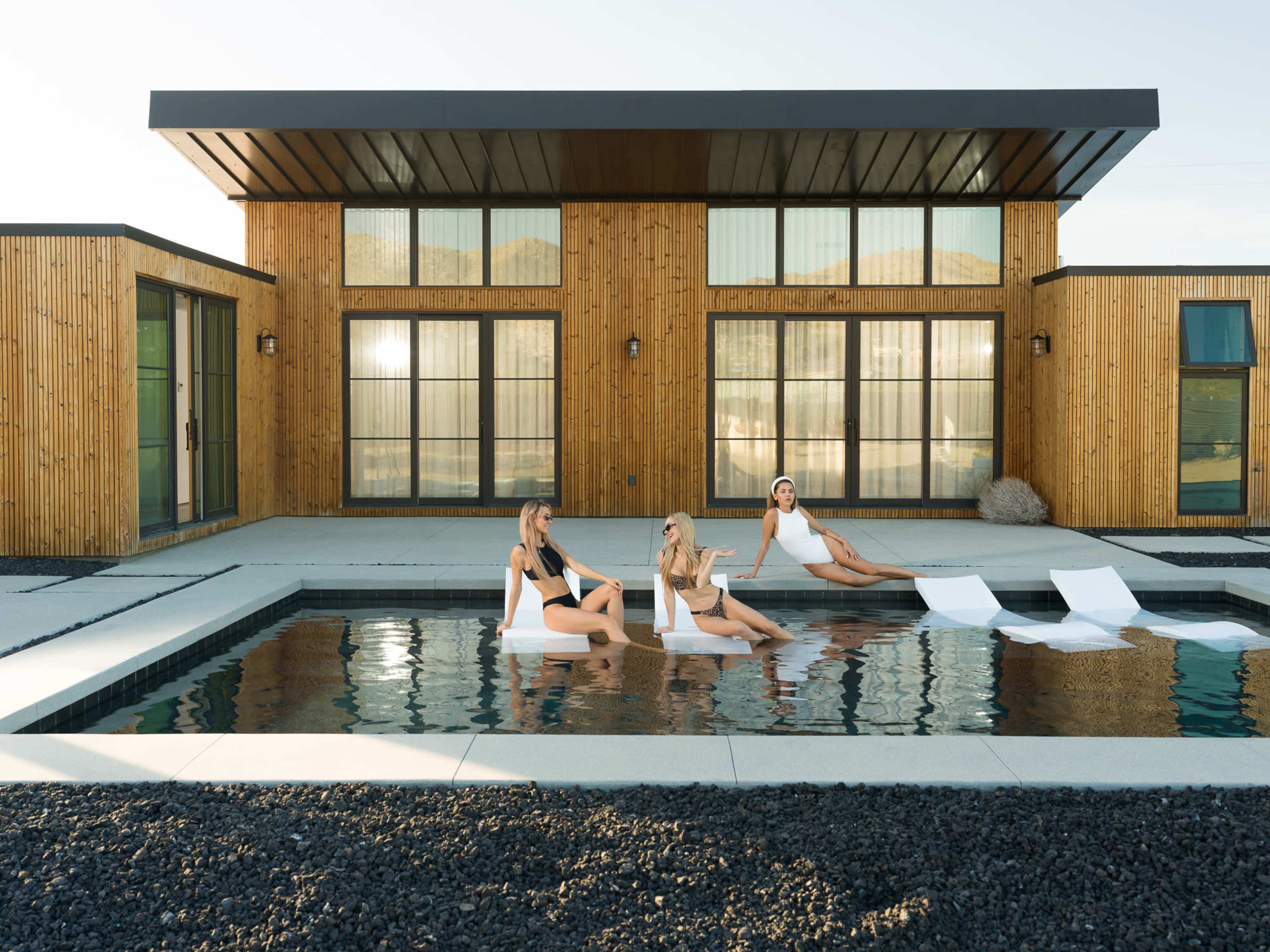 Three women relax on floating loungers in a modern pool in front of a stylish wooden-facade house.