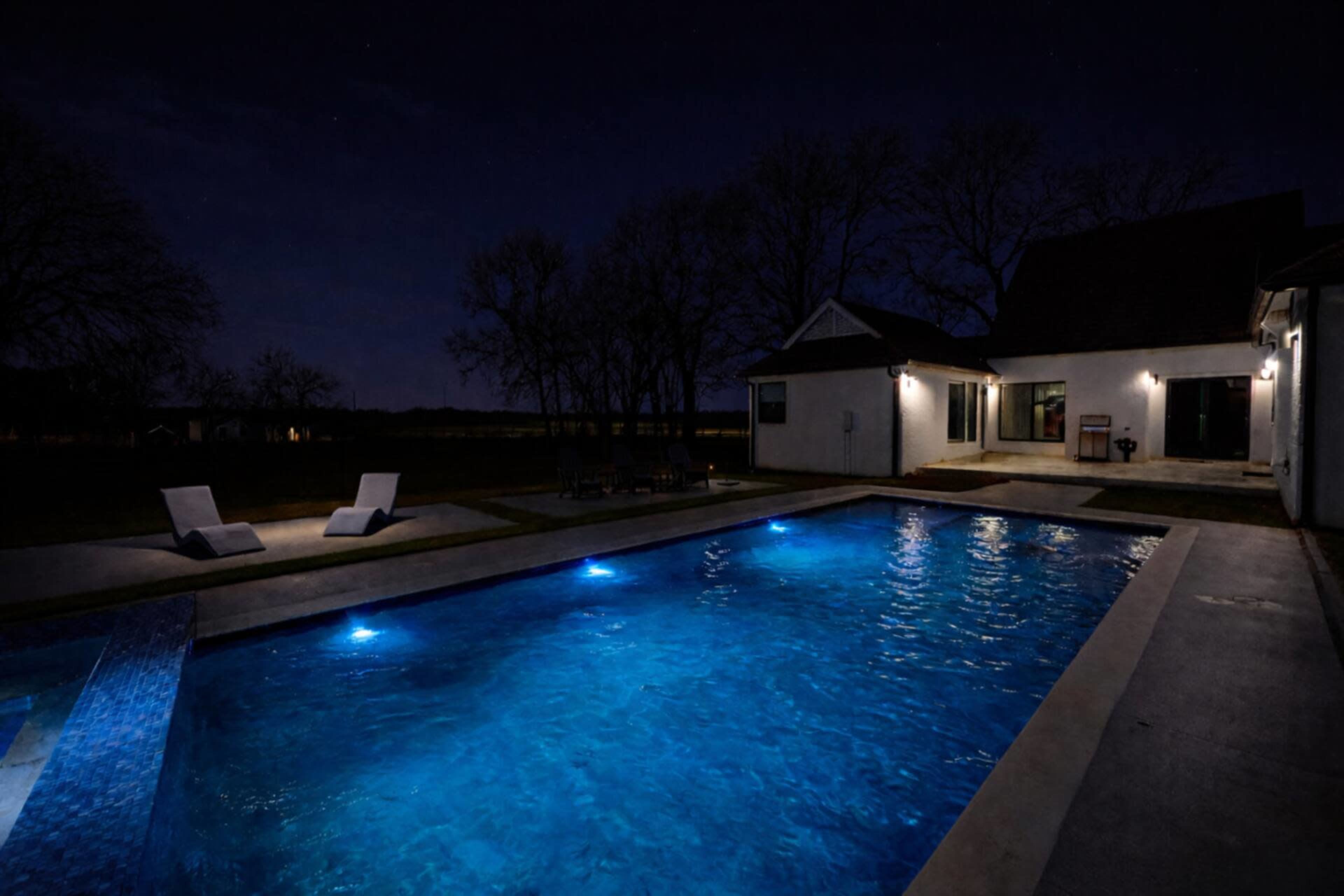 A well-lit swimming pool is surrounded by a house and lawn under a night sky.