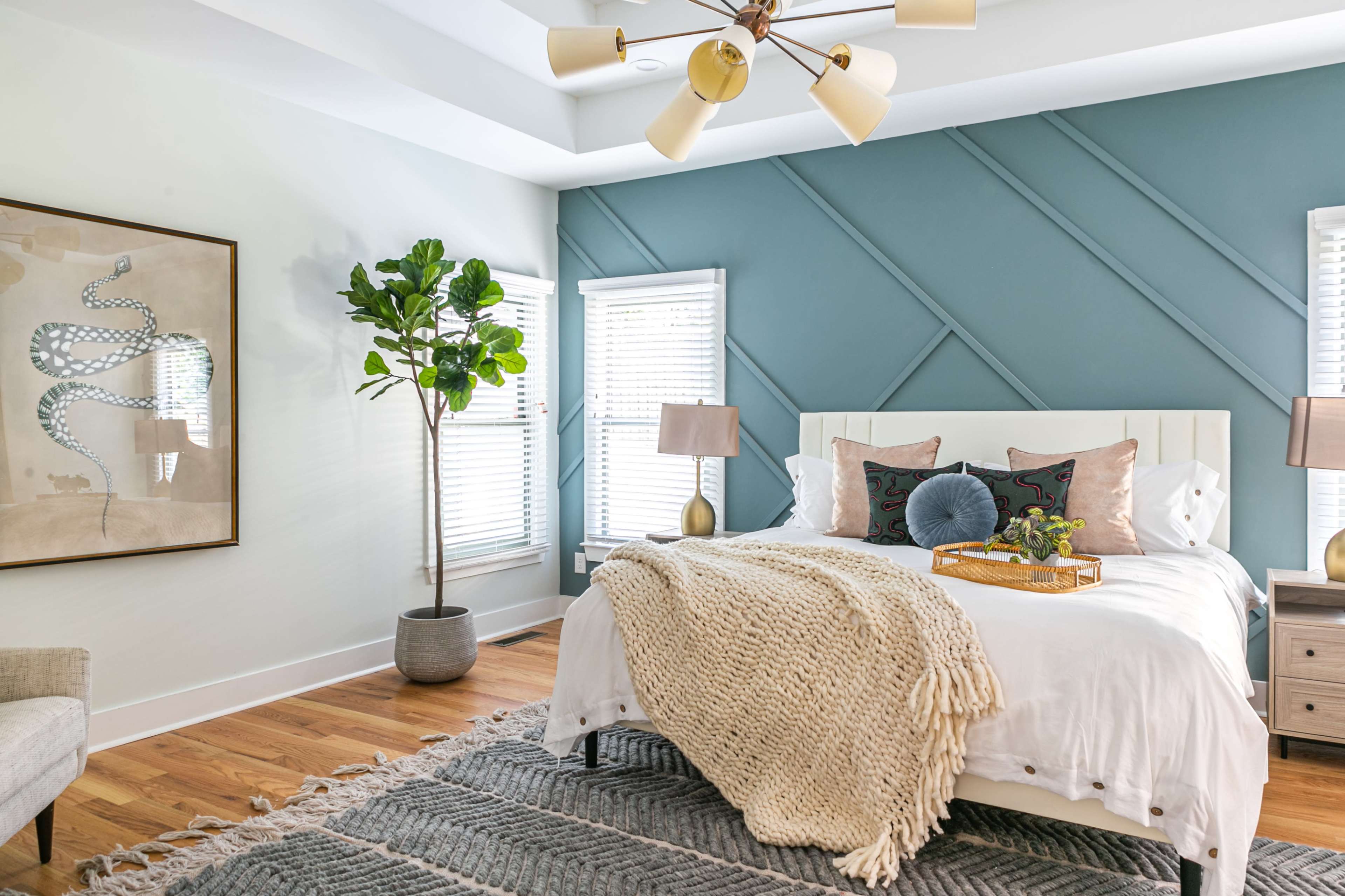 A well-decorated bedroom featuring a white bed with decorative pillows, a large wall art piece, and a potted plant beside windows.