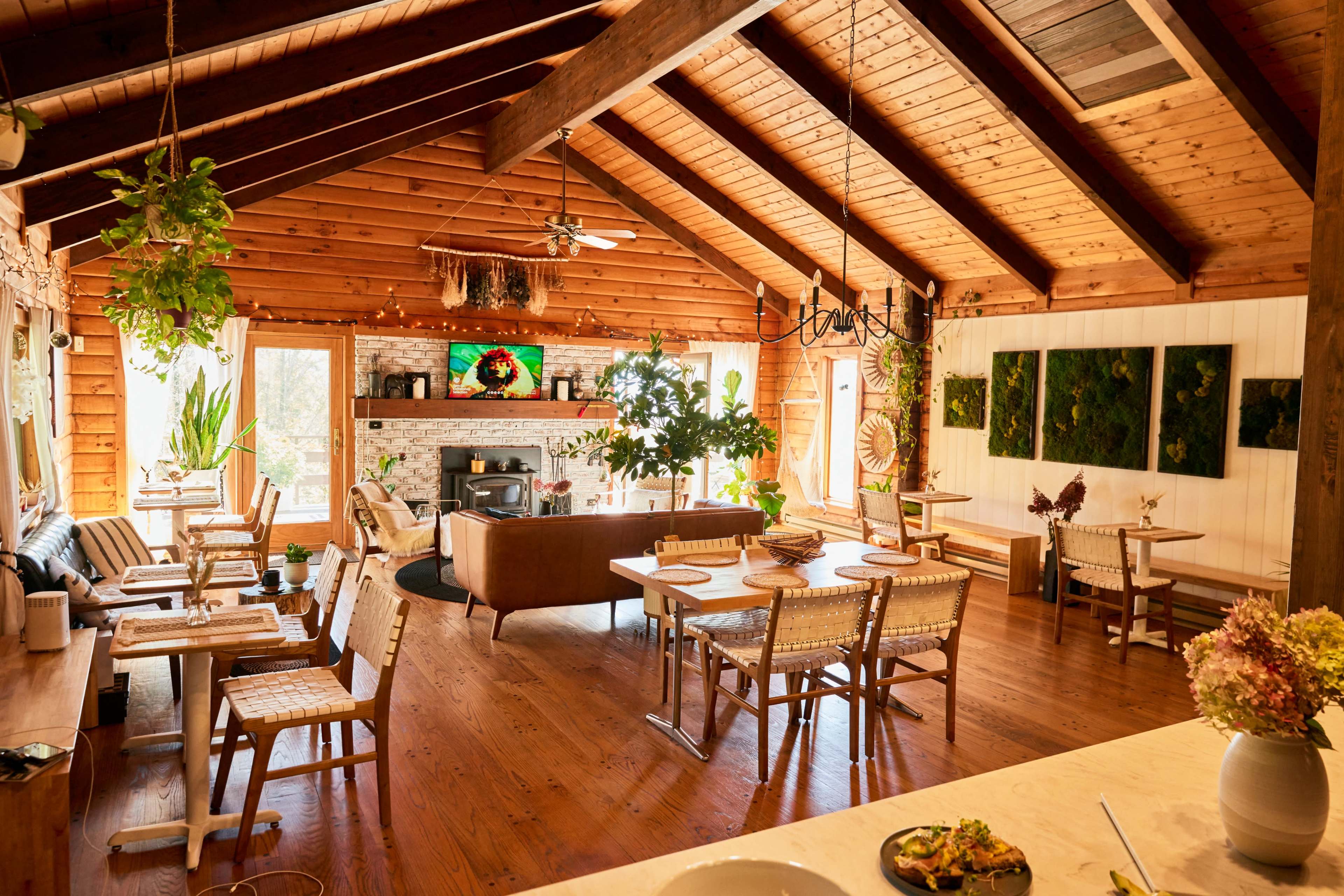 A spacious wooden interior features a dining area with several chairs and tables, a living space with a fireplace, and large windows allowing natural light.